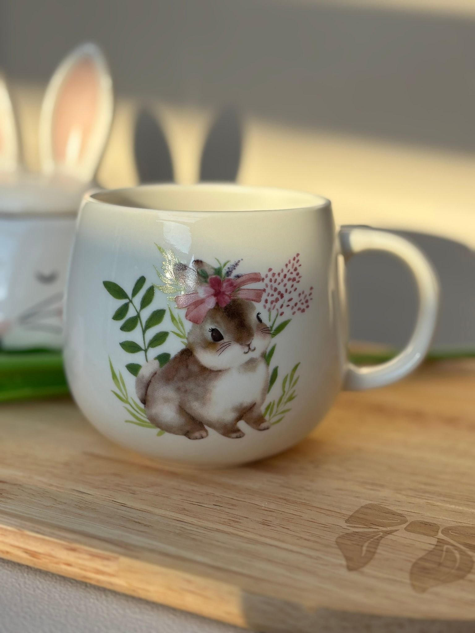 Een witte keramische mok met een geschilderde afbeelding van een konijntje met een roze bloemenkrans, zittend op een houten ondergrond.
