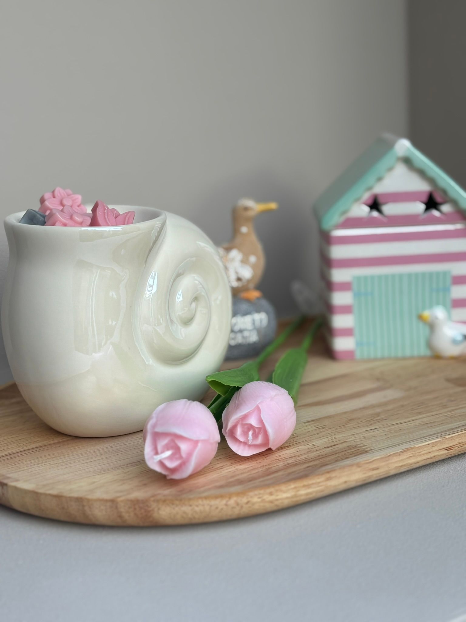 Een crèmekleurige, slakvormige kaarsenhouder gevuld met roze bloemvormige geurblokjes, naast twee roze tulpvormige kaarsen op een dienblad.