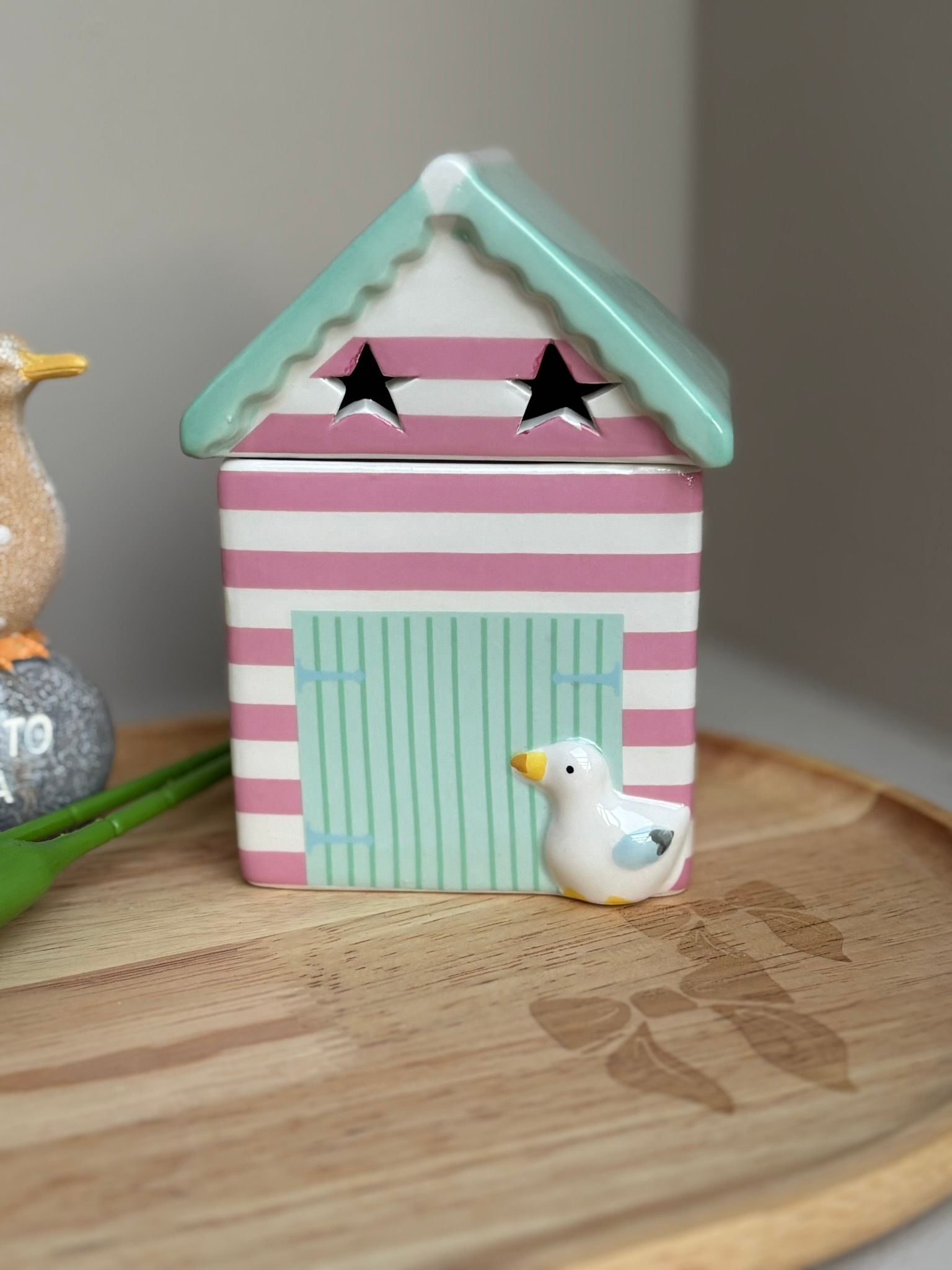 Een decoratief keramisch strandhuisje met roze en witte strepen, een turquoise dak met stervormige uitsparingen en een klein meeuwenbeeldje.