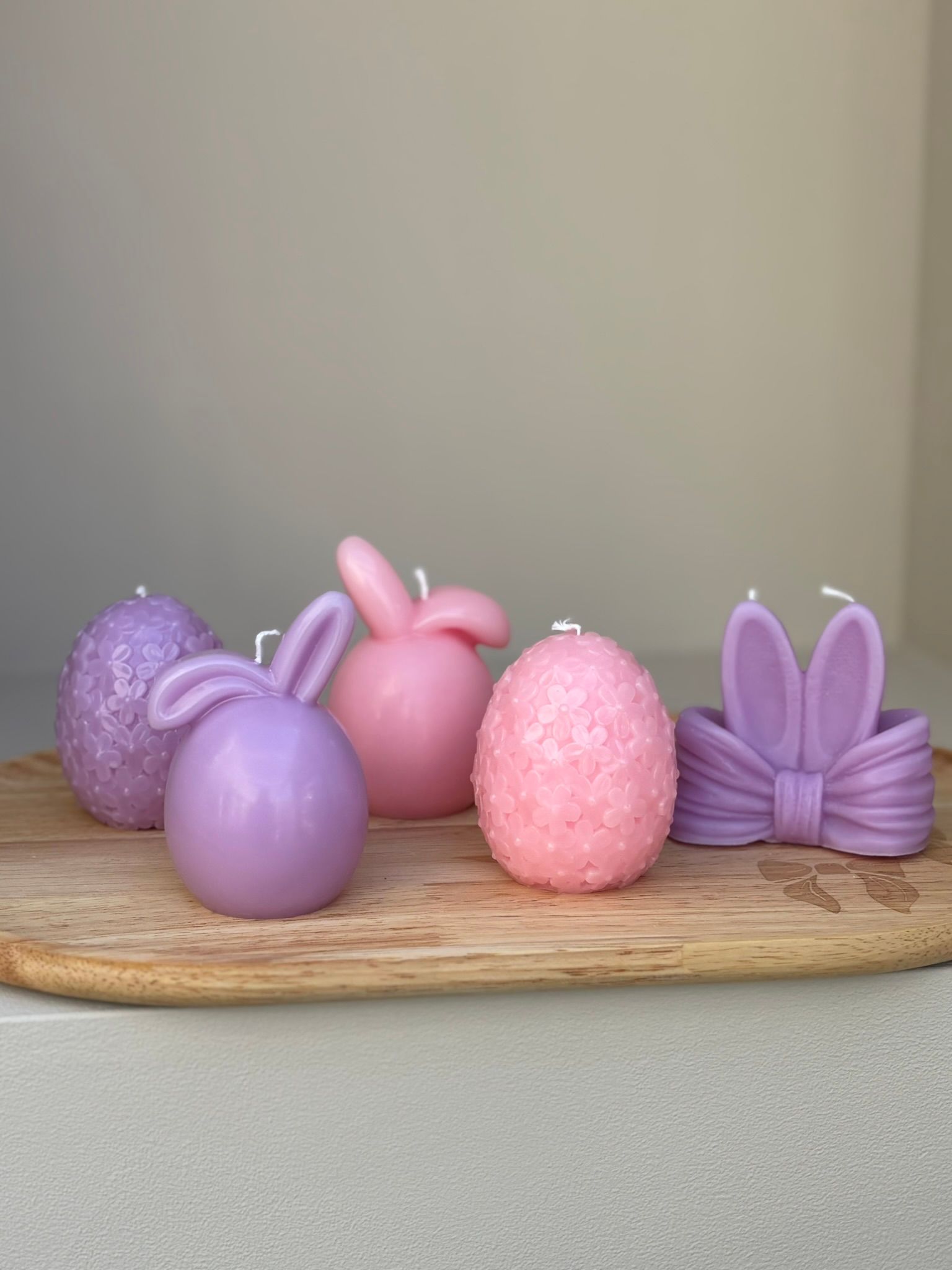 Drie paasdecoraties op een houten plank: een beige pluche konijntje, een roze eivormige kaars met bloemenmotief en een paars paasei.