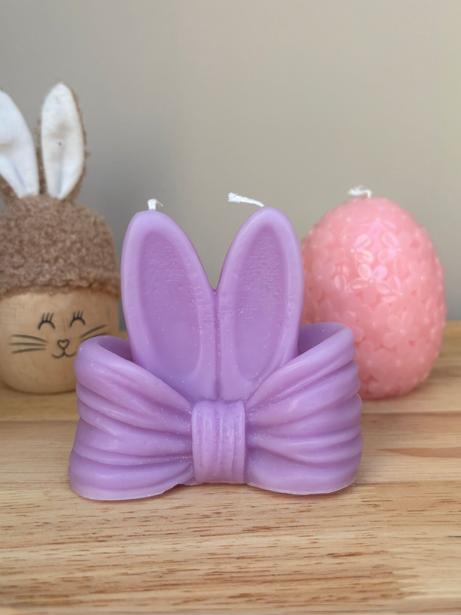 Een paarse, strikvormige kaars met konijnenoortjes staat op een houten dienblad naast een ei met konijnenoortjes en een roze ei met een structuur.