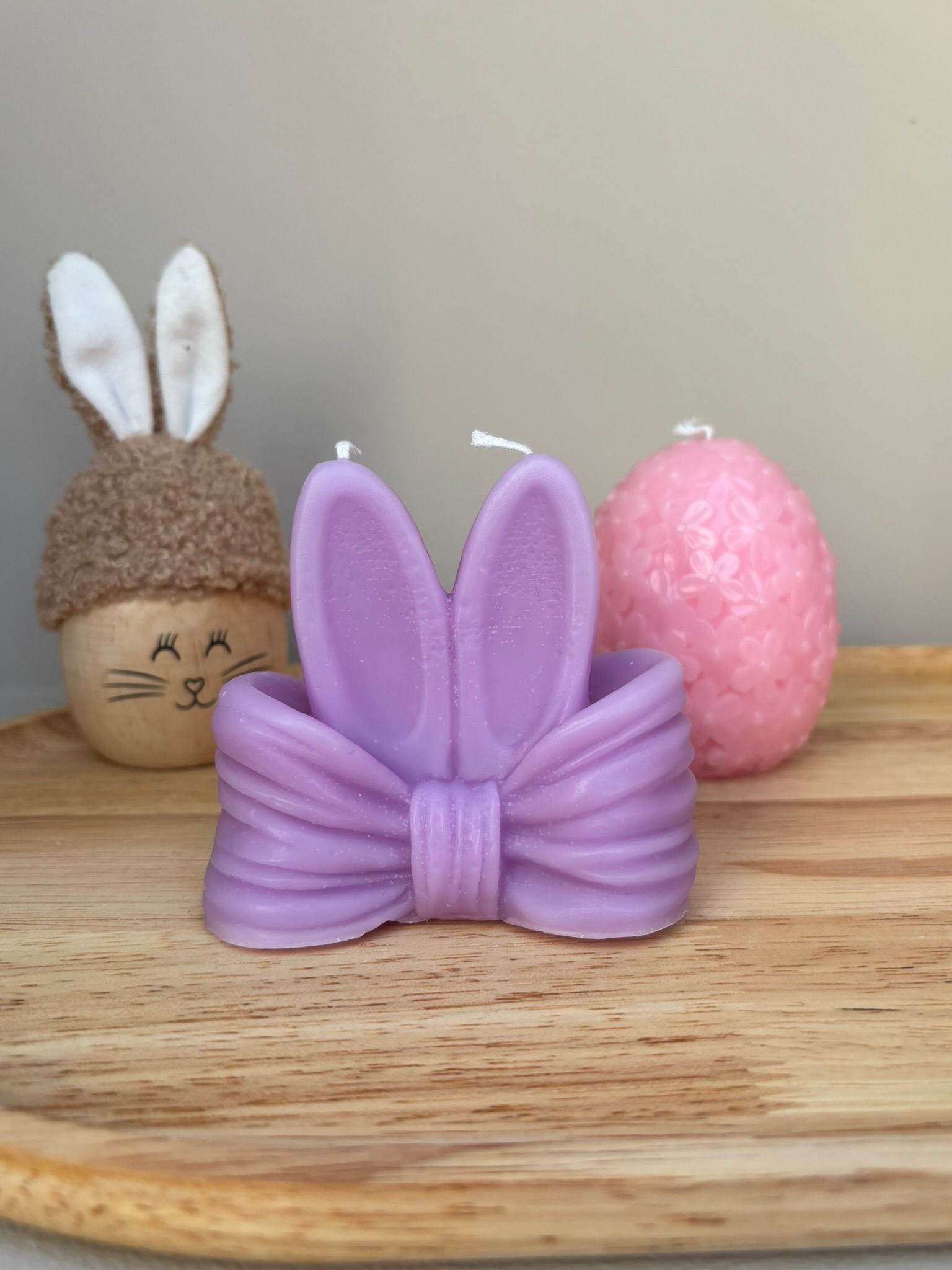 Een paarse, strikvormige kaars met konijnenoortjes staat op een houten dienblad naast een ei met konijnenoortjes en een roze ei met een structuur.