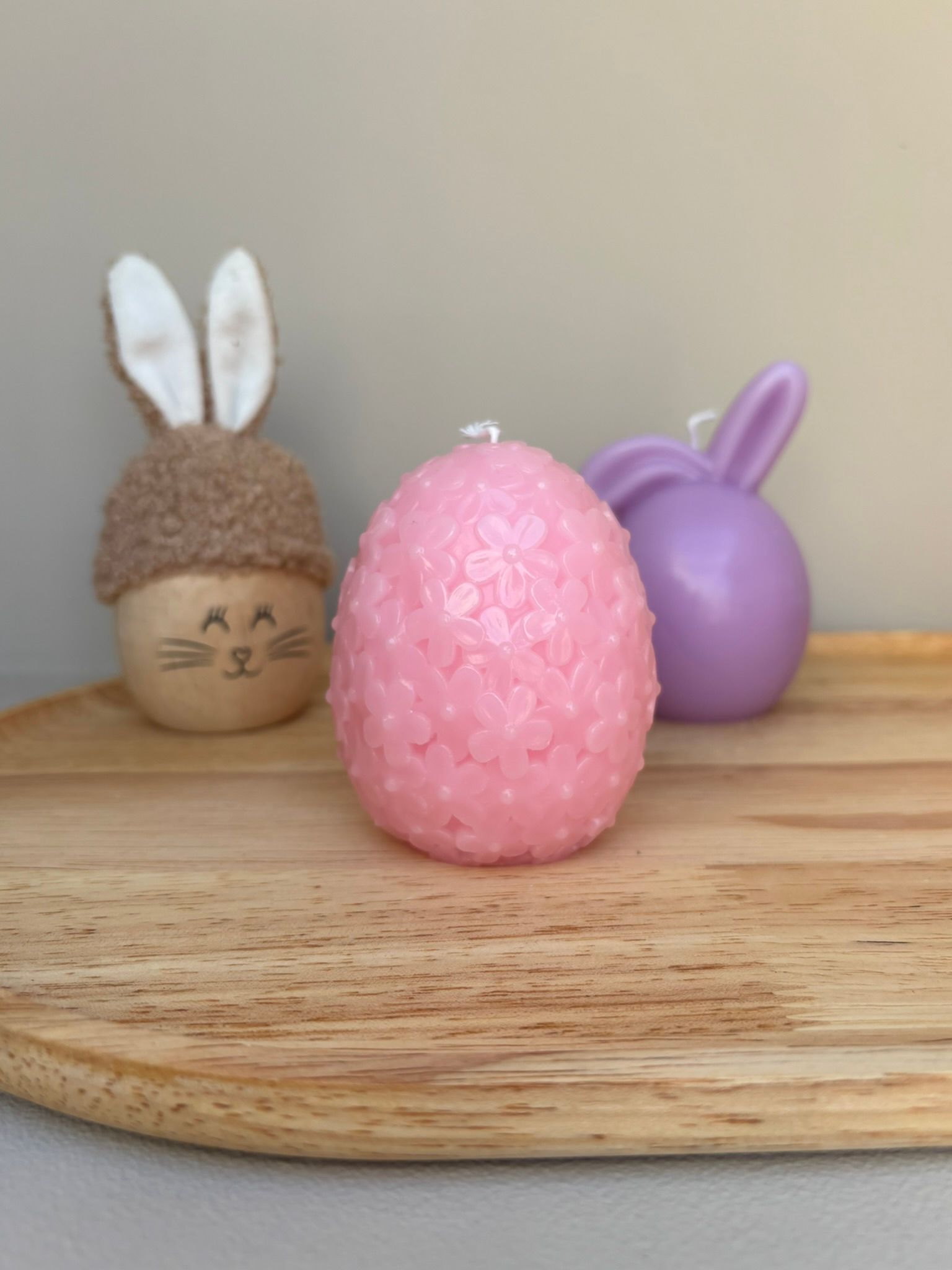 Drie paasdecoraties op een houten plank: een beige pluche konijntje, een roze eivormige kaars met bloemenmotief en een paars paasei.