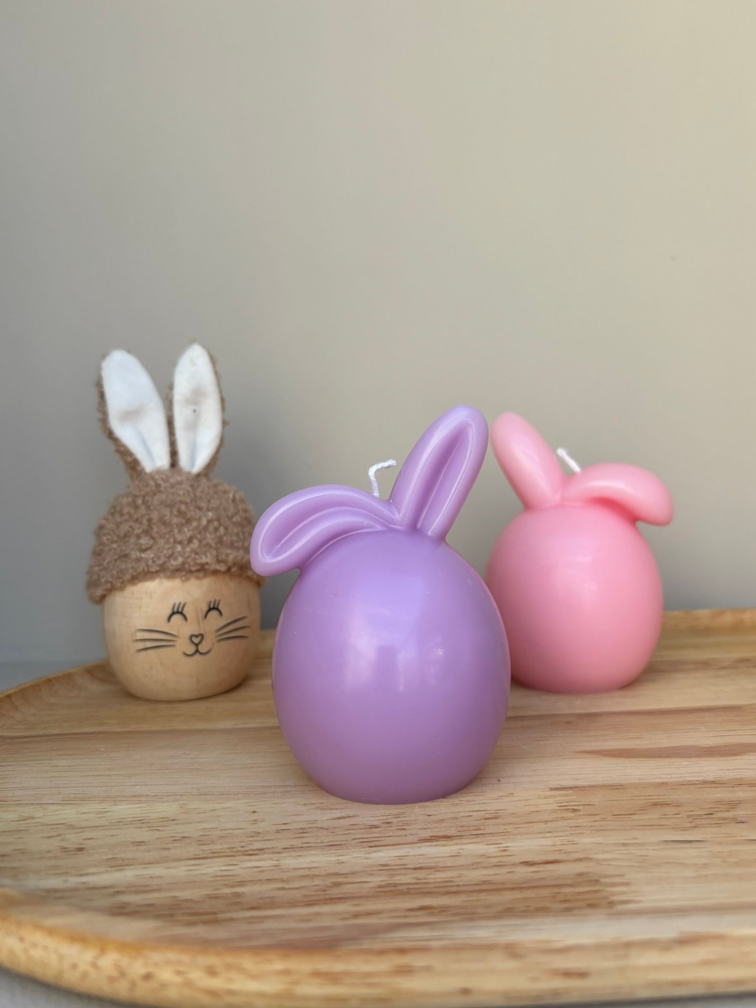 Drie decoraties met konijntjesthema: een konijntje van vilt, een paarse kaars en een roze kaars op een houten dienblad.