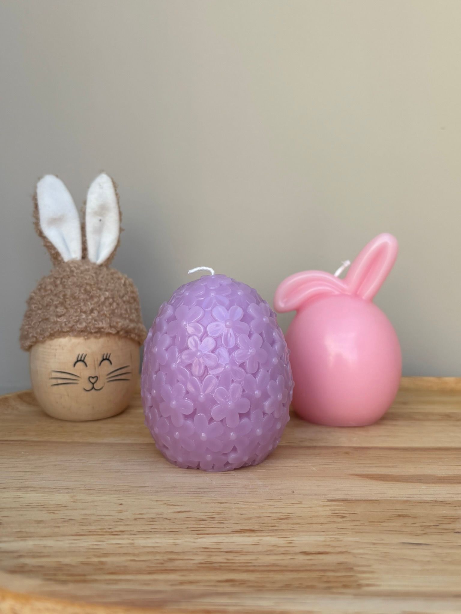 Drie paasdecoraties op een houten dienblad: een paasei met konijnenoortjes, een paars paasei met bloemen en een roze paasei met een konijntje.