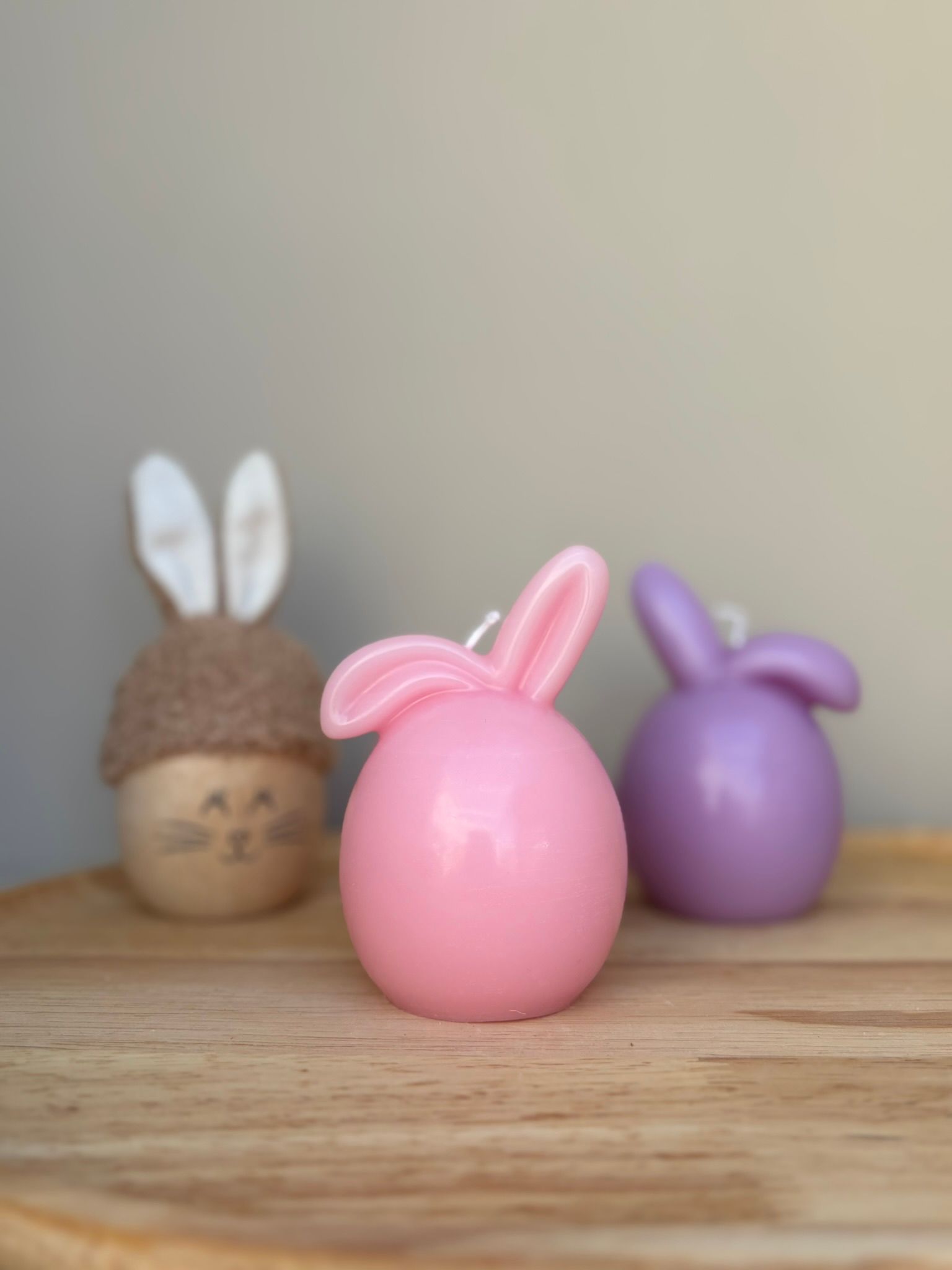 Drie objecten met een konijnenthema op een houten ondergrond: een bruin stoffen konijn, een roze kaars en een paarse kaars.