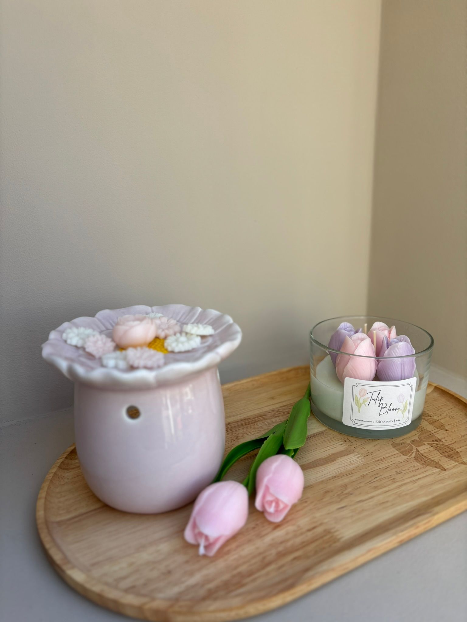 Een decoratieve kaars in een glazen pot, afgedekt met roze en paarse tulpvormige was, staat op een houten dienblad met twee tulpen.