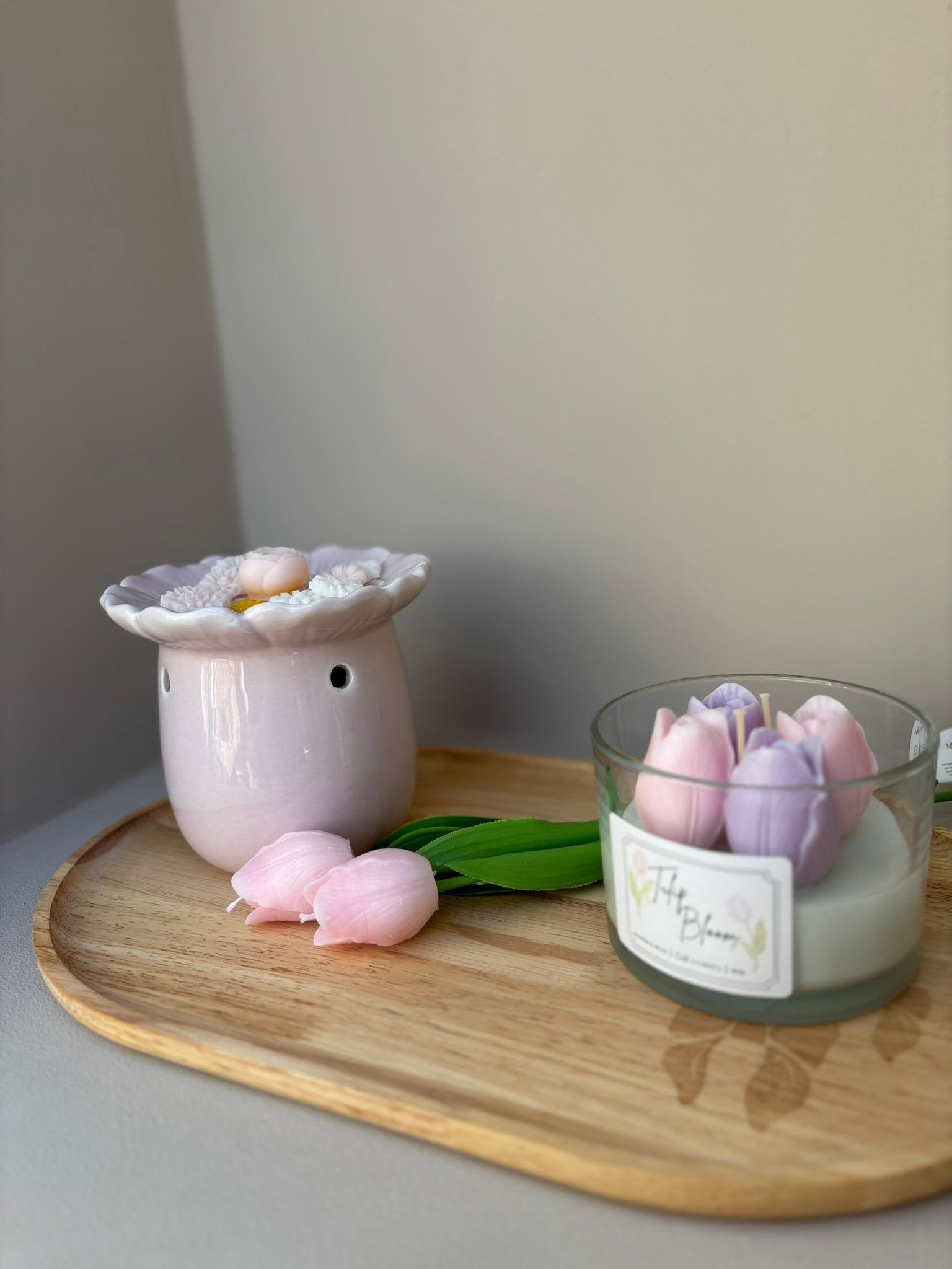 Een lichtroze keramische kaarsenwarmer in de vorm van een madeliefje, rustend op een houten schaaltje met twee kleine roze bloemknopjes.