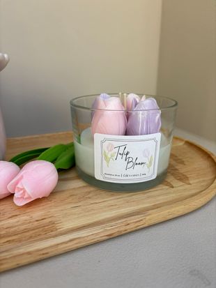 Een glazen kaarsenpot met daarop roze en paarse tulpvormige wasstukjes, die op een houten dienblad met twee roze tulpen staat.