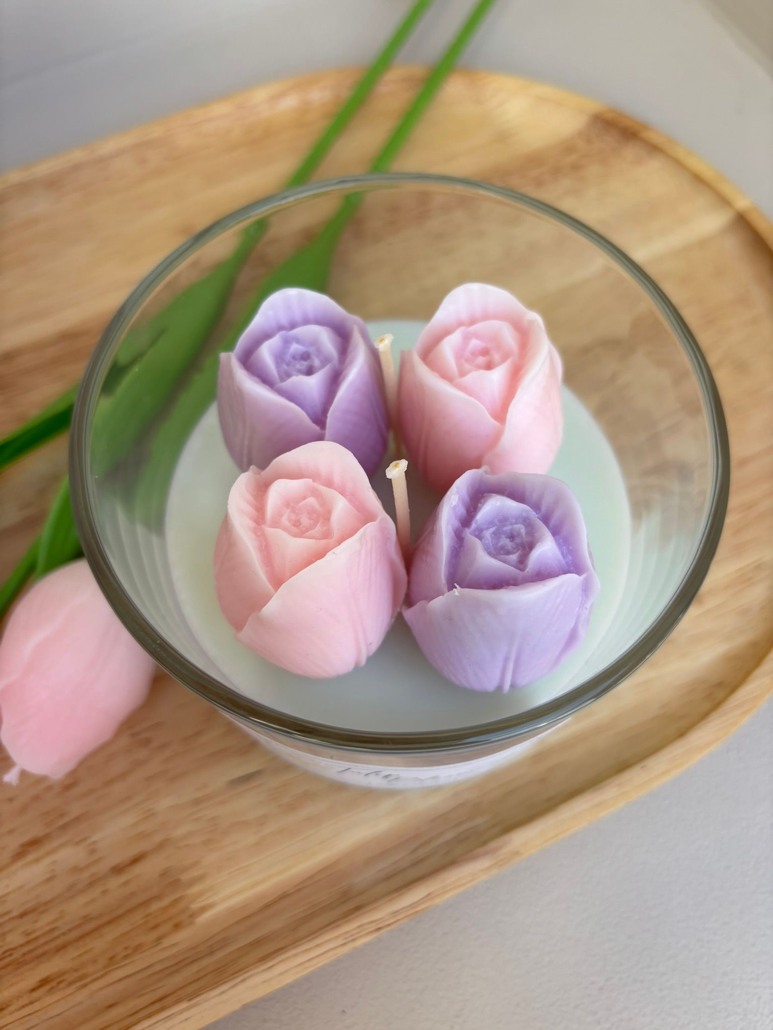 Vier pastelroze en paarse, roosvormige kaarsknopjes, gerangschikt in een glazen schaal op een houten dienblad.