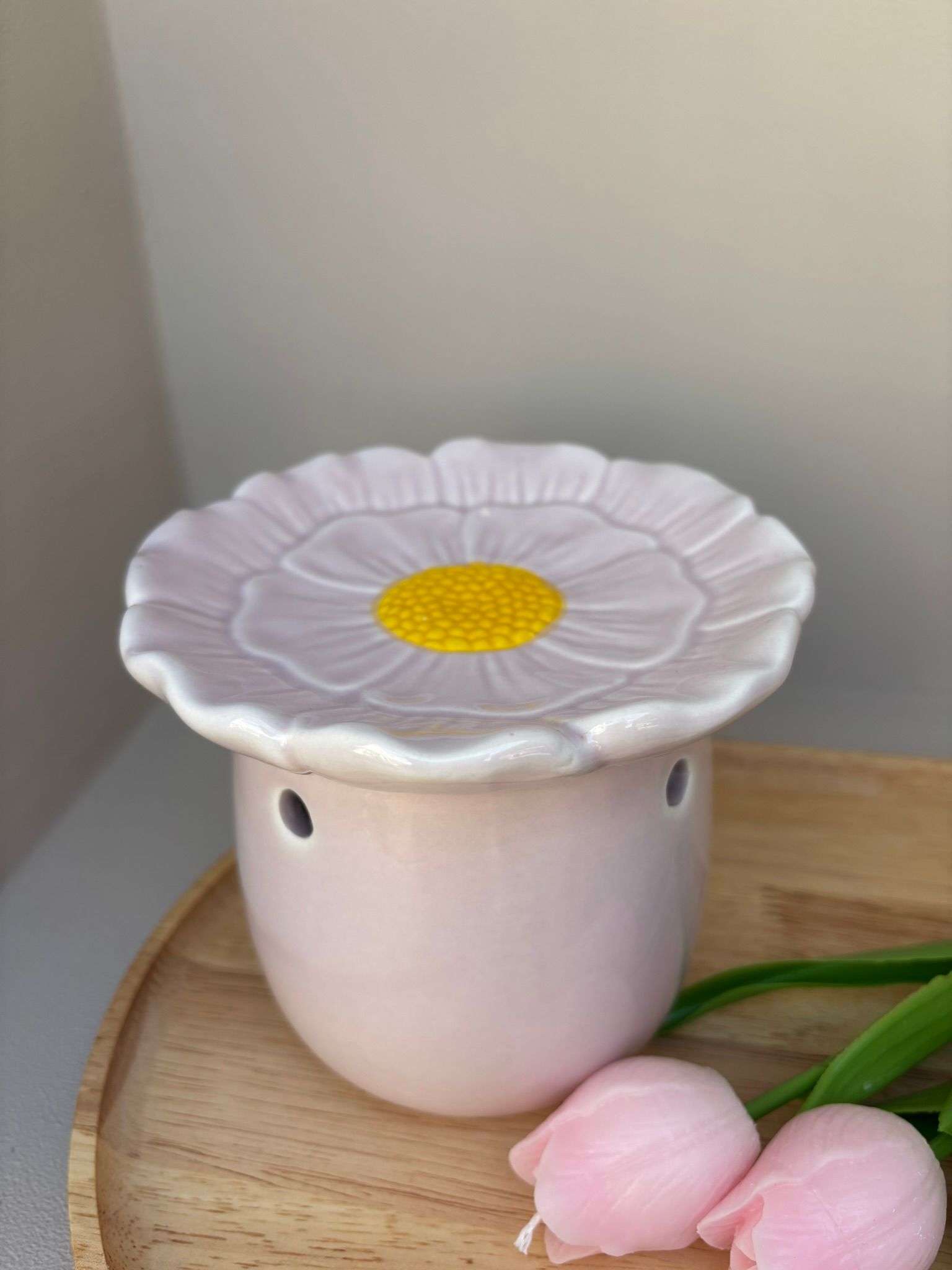 Een lichtroze keramische kaarsenwarmer in de vorm van een madeliefje, rustend op een houten schaaltje met twee kleine roze bloemknopjes.