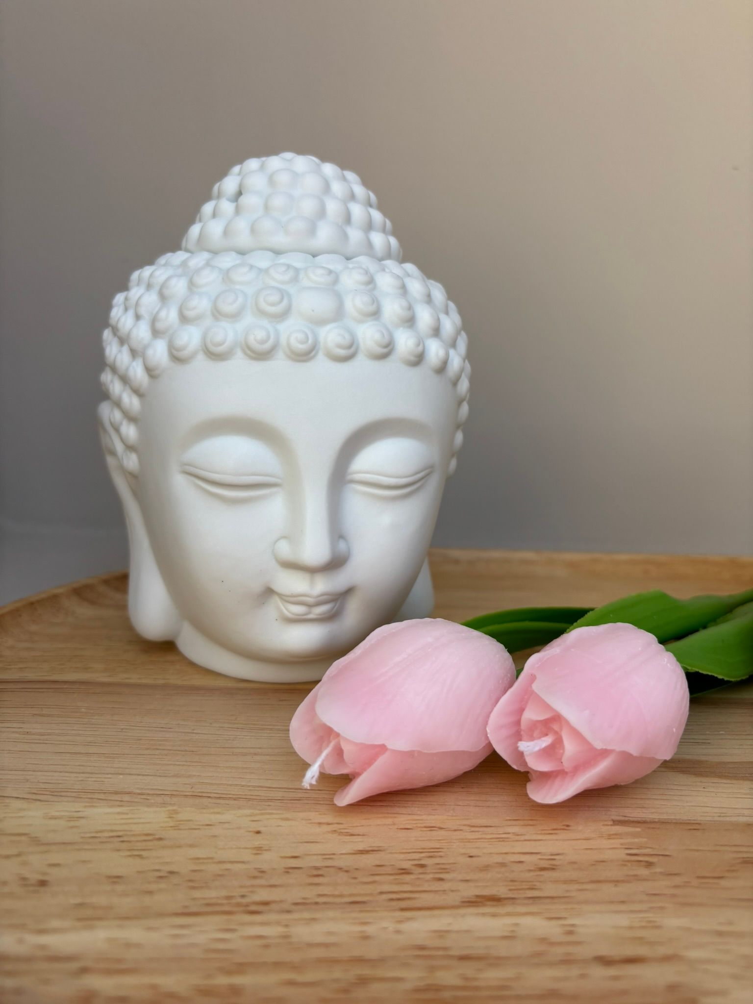 Een wit Boeddhabeeldje staat op een houten dienblad naast twee kleine, lichtroze, tulpvormige kaarsen.