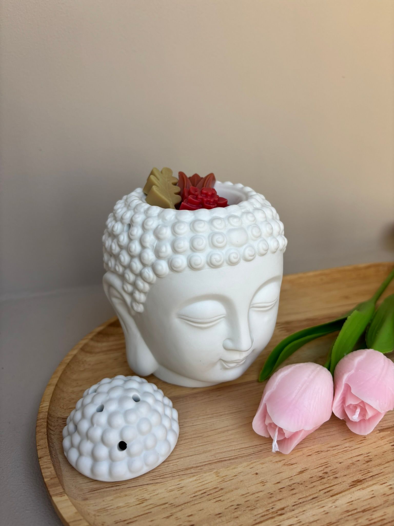 Een wit Boeddhabeeldje staat op een houten dienblad naast twee kleine, lichtroze, tulpvormige kaarsen.