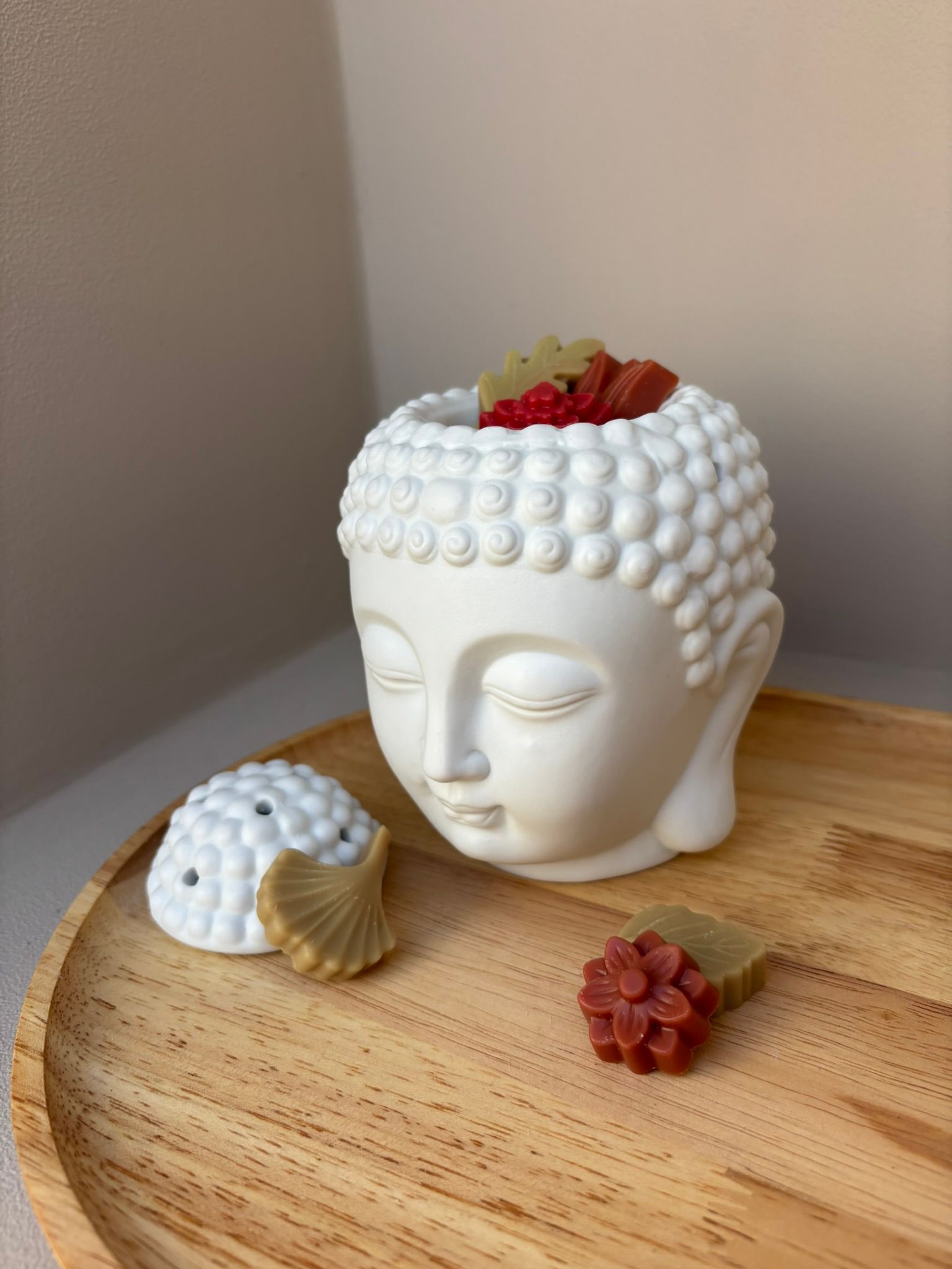 Een klein hoopje bloemvormige was, in rode en beige tinten, smelt op een houten ondergrond voor een wit Boeddhabeeld.