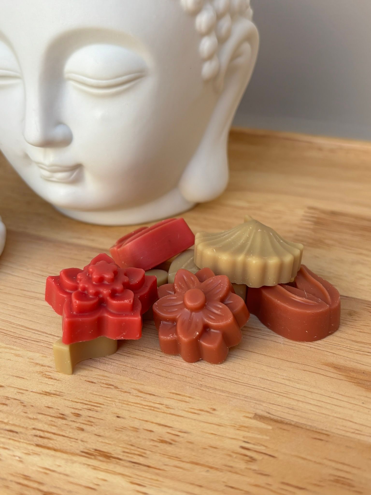 Een klein hoopje bloemvormige was, in rode en beige tinten, smelt op een houten ondergrond voor een wit Boeddhabeeld.