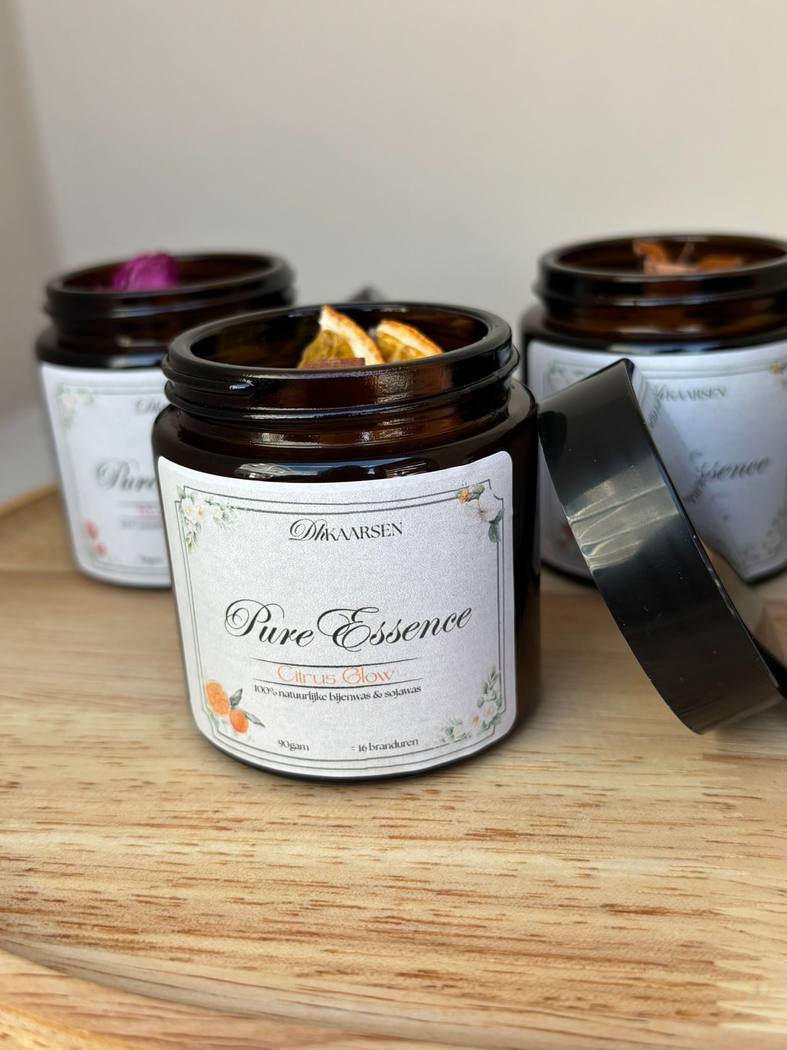 Drie amberkleurige glazen kaarsenhouders met etiketten en gedroogde sinaasappelschijfjes op een houten ondergrond, één houder met deksel ernaast.