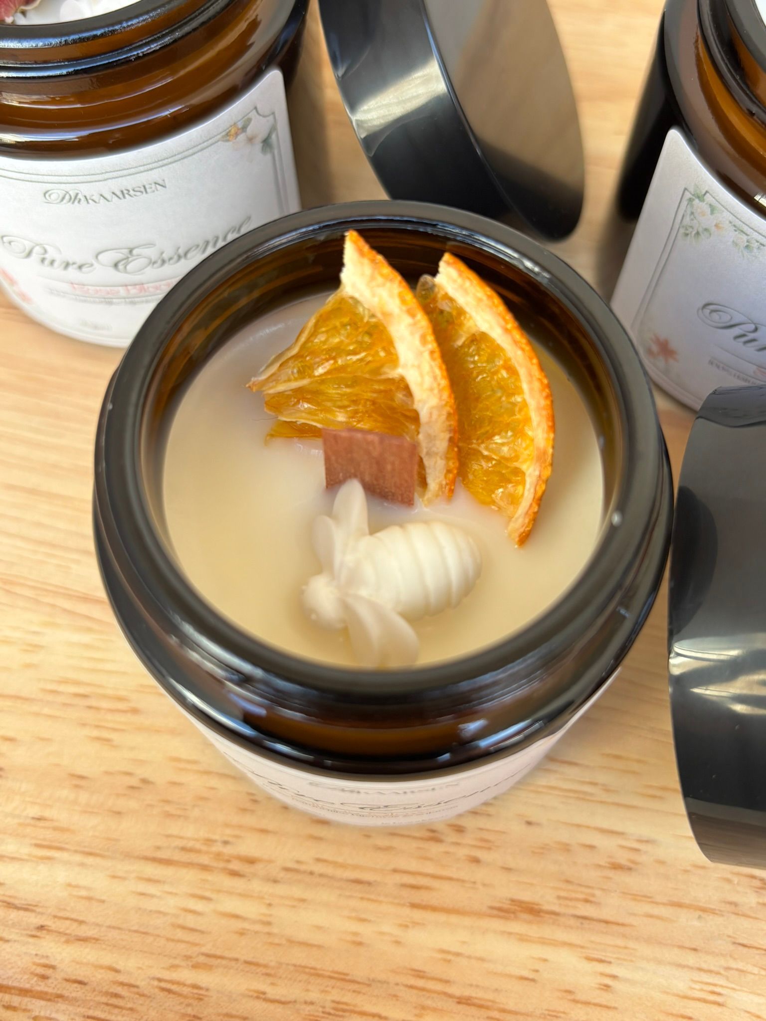 Drie amberkleurige glazen kaarsenhouders met etiketten en gedroogde sinaasappelschijfjes op een houten ondergrond, één houder met deksel ernaast.