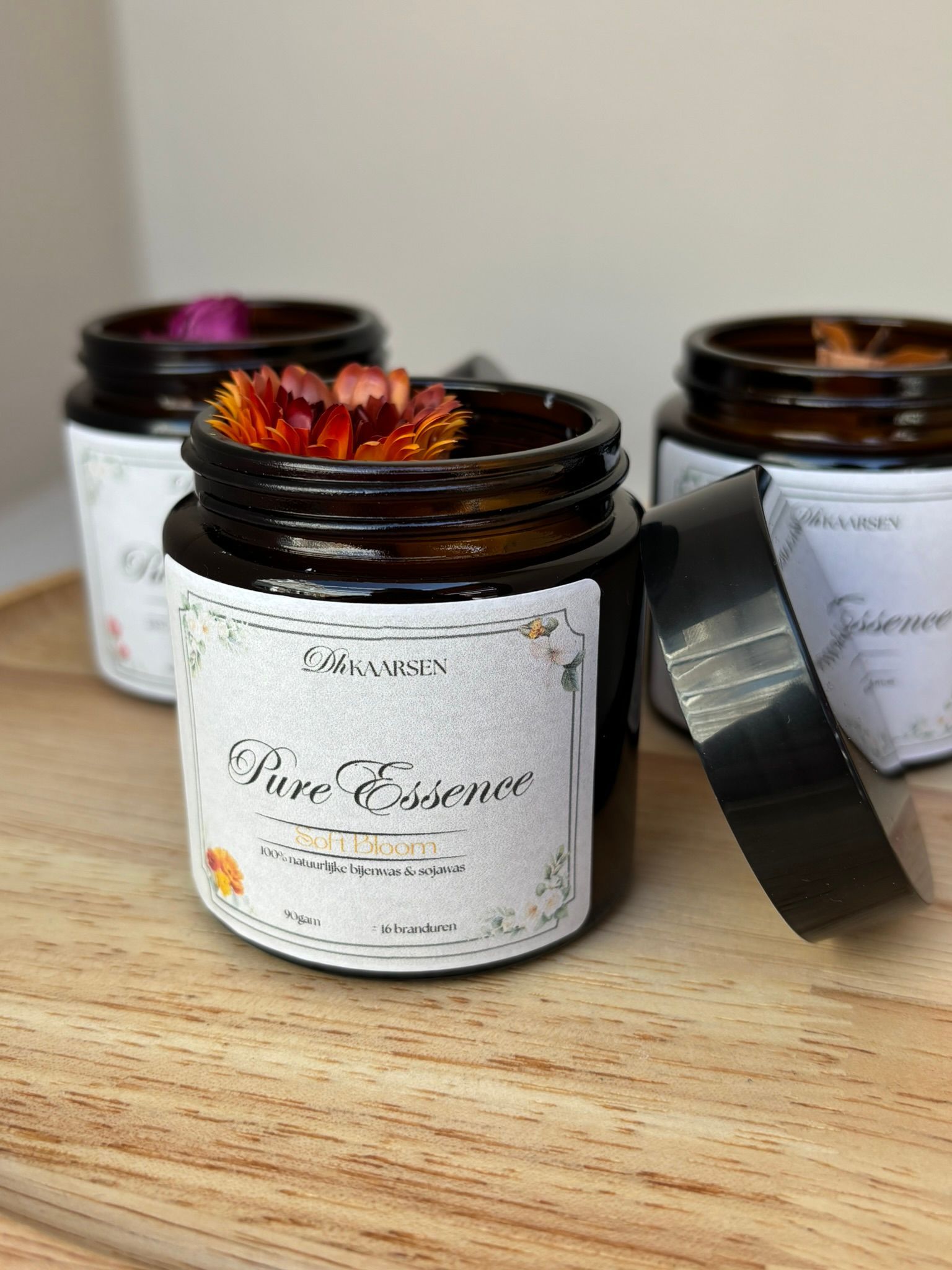 Drie amberkleurige glazen potjes met witte etiketten en bloemversieringen staan ​​op een houten ondergrond, waarvan er één het deksel heeft verwijderd.