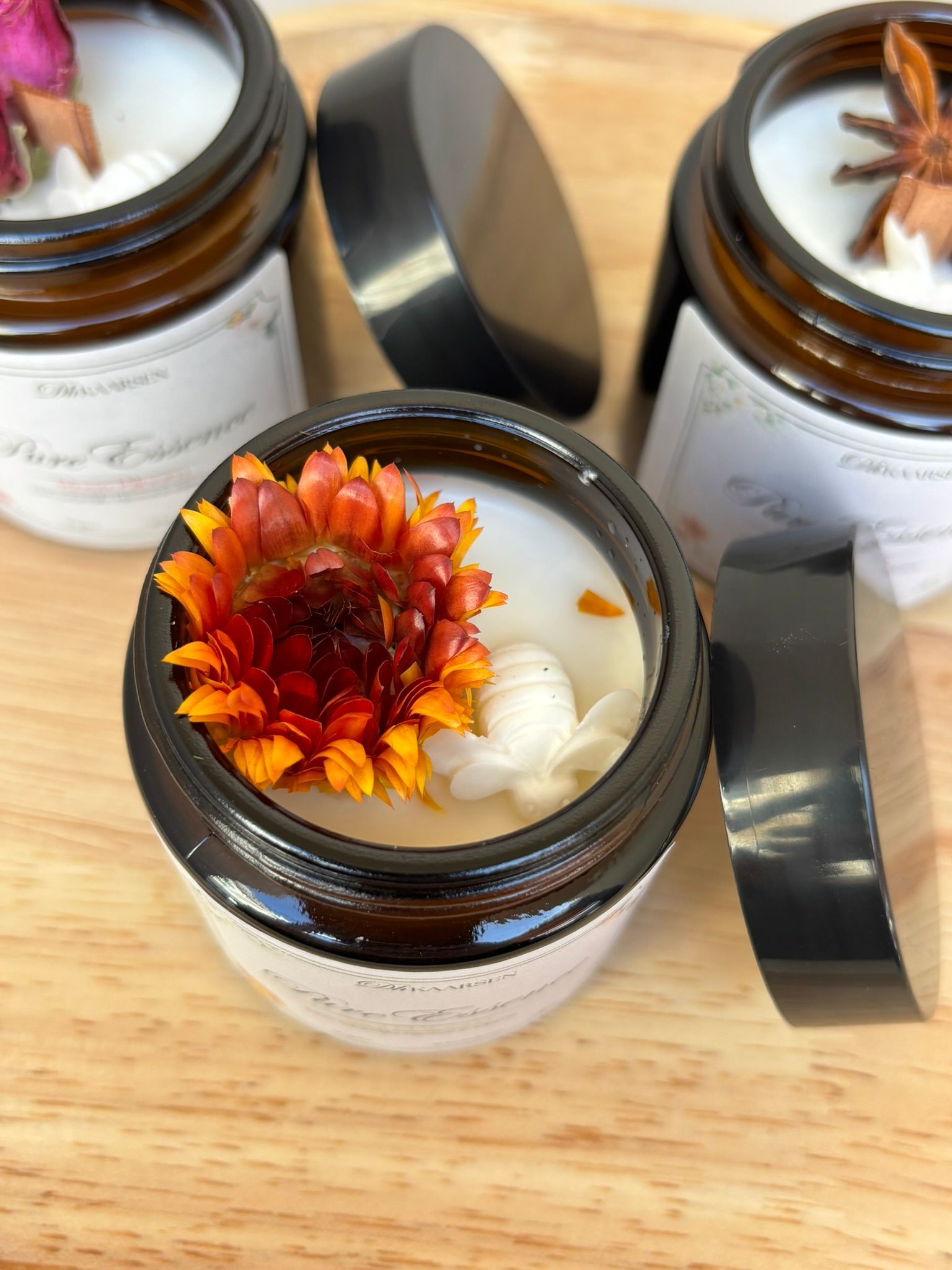Drie amberkleurige glazen potjes met witte etiketten en bloemversieringen staan ​​op een houten ondergrond, waarvan er één het deksel heeft verwijderd.