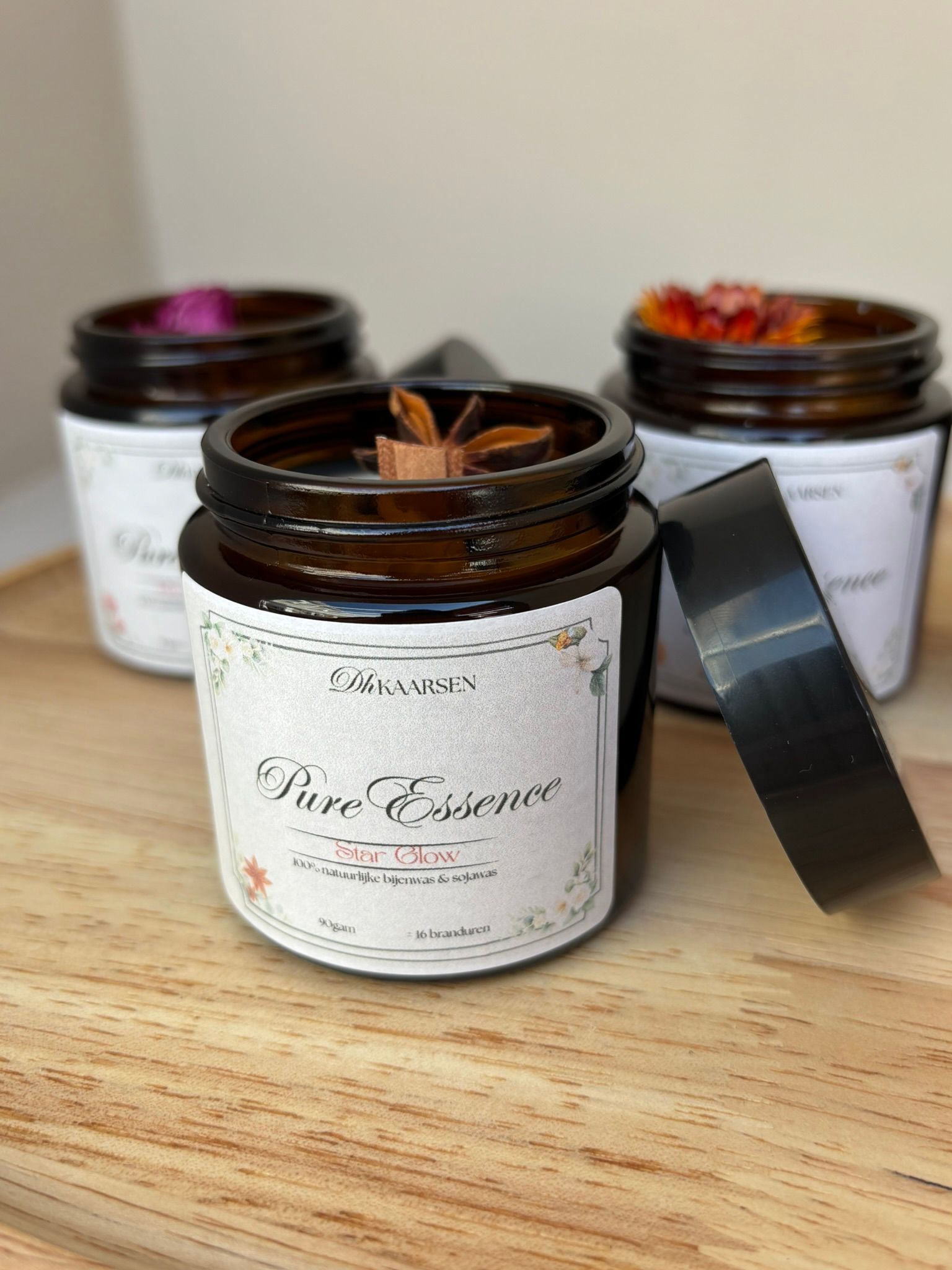 Drie amberkleurige glazen potjes gevuld met kaarsen op een houten ondergrond, elk versierd met een gedroogde bloem of steranijs als decoratie.