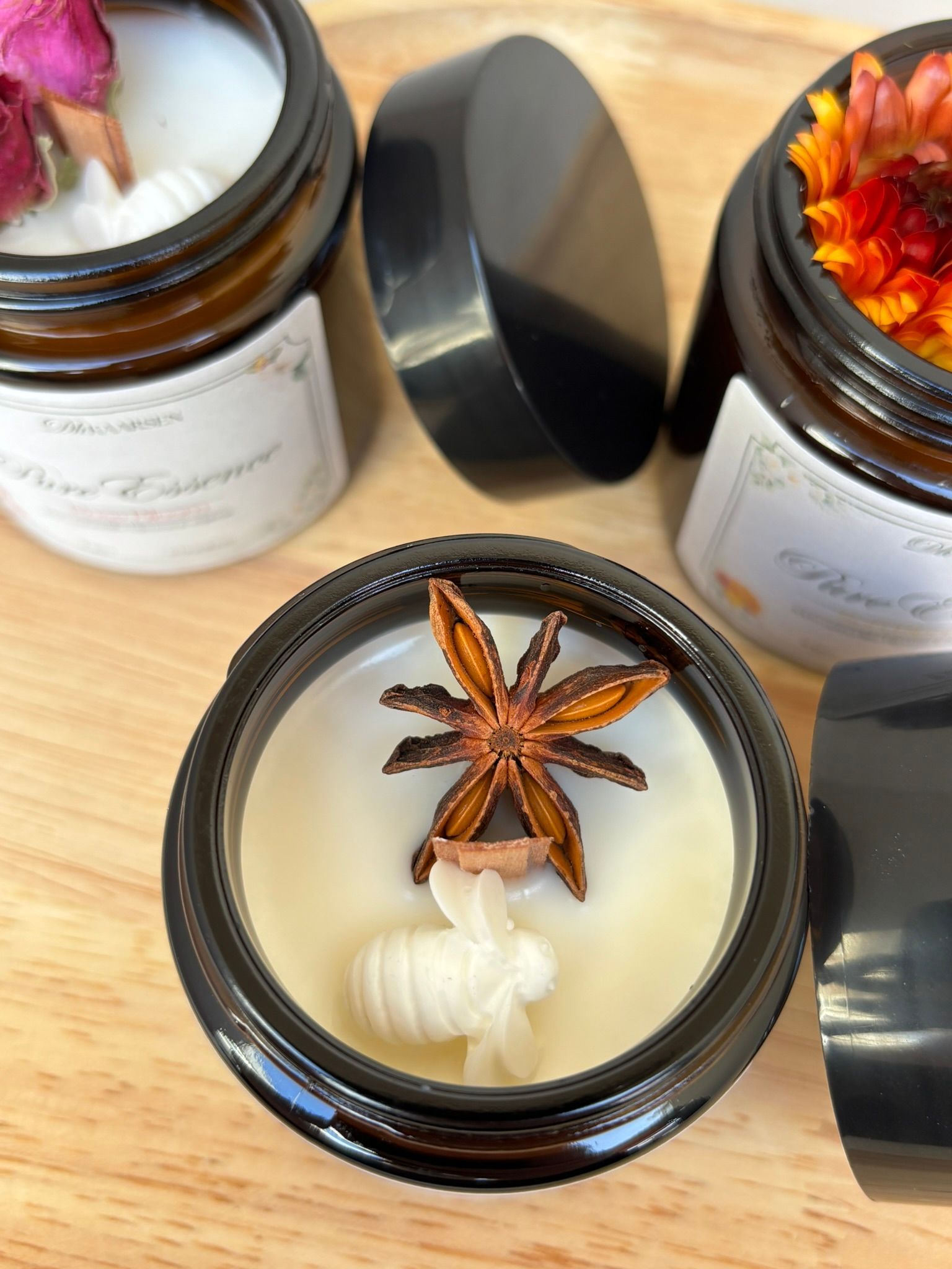 Drie amberkleurige glazen potjes gevuld met kaarsen op een houten ondergrond, elk versierd met een gedroogde bloem of steranijs als decoratie.