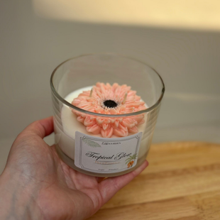 Hand die een glazen pot met kaars vasthoudt, met een roze bloem erop en een decoratief etiket.