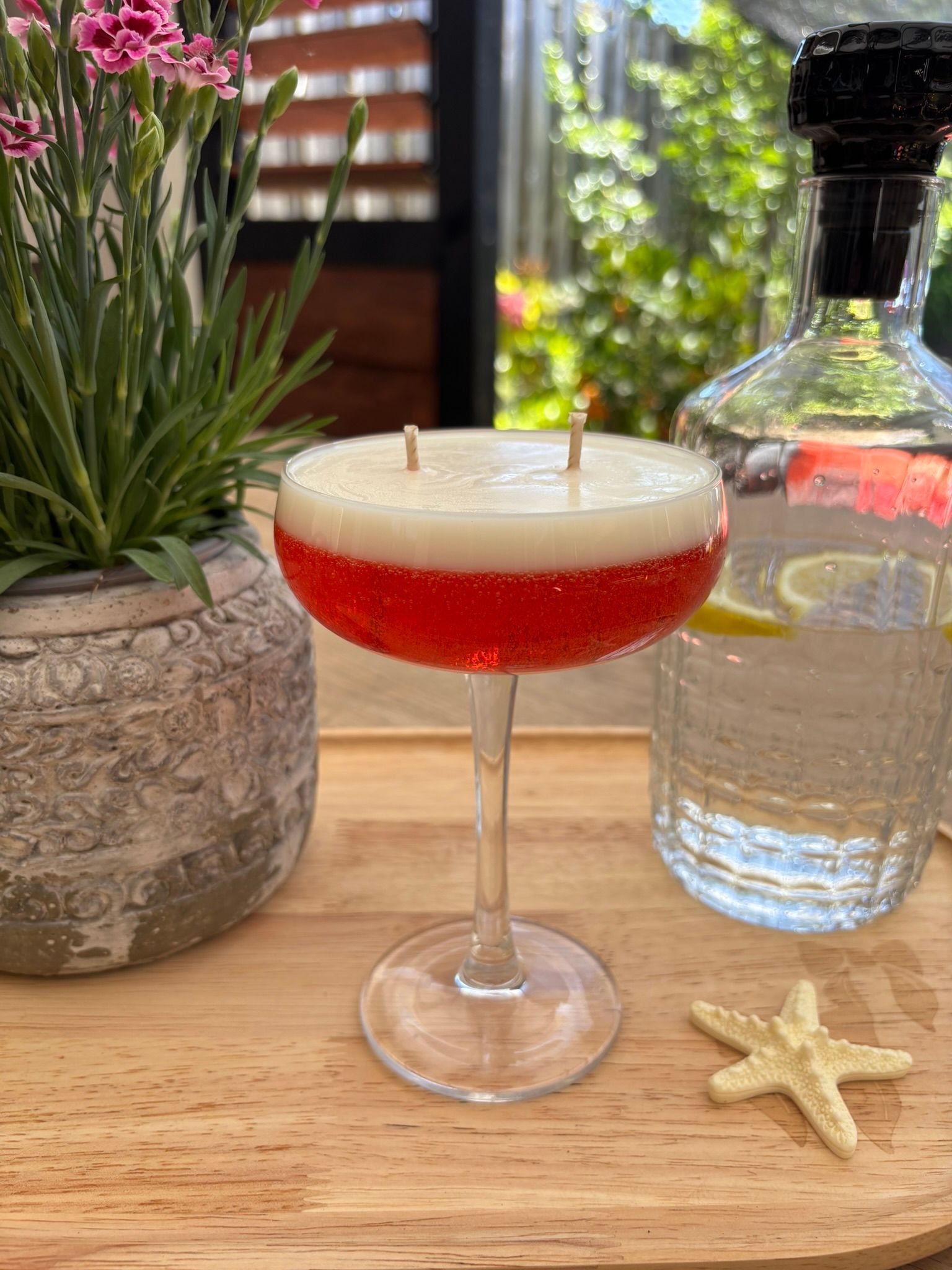 Een rode cocktail in een coupeglas naast een fles water, een potplant en een zeester op een houten tafel.