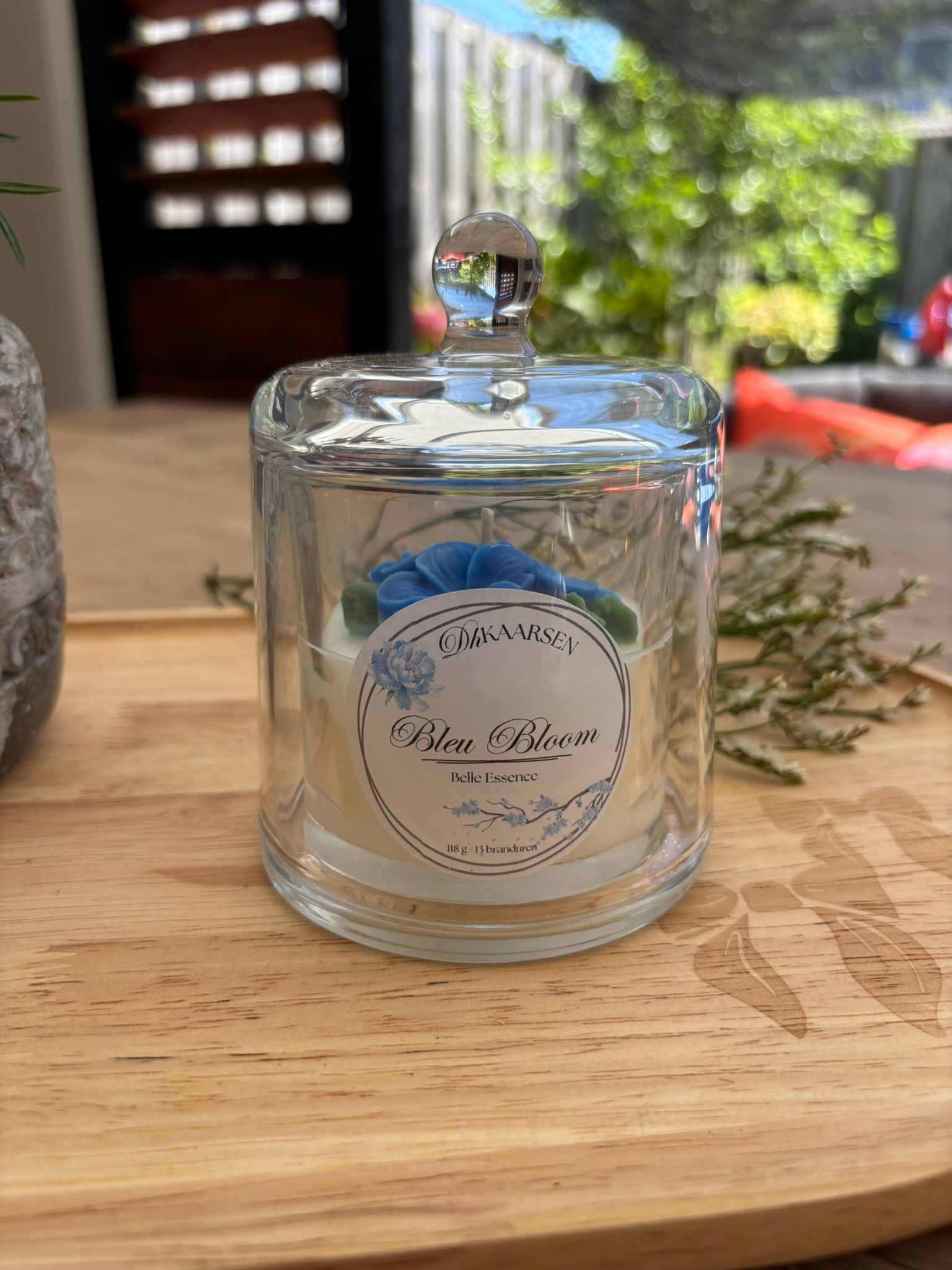 Een kaars in een glazen pot met een blauw etiket staat op een houten tafel buiten, naast een glazen stolp.