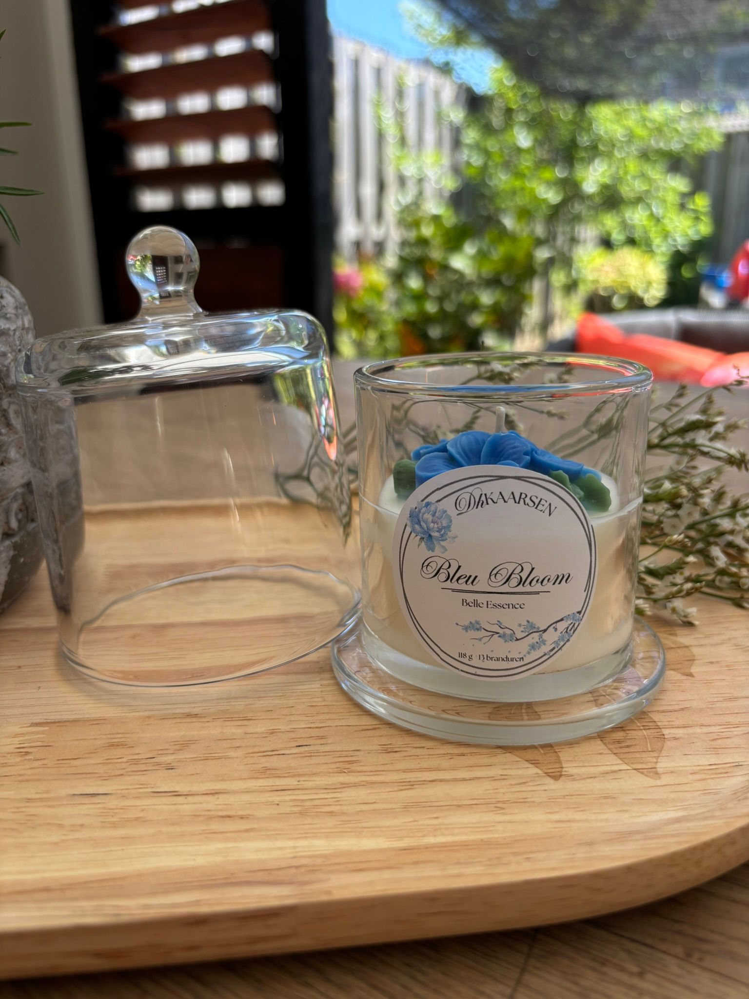 Een kaars in een glazen pot met een blauw etiket staat op een houten tafel buiten, naast een glazen stolp.