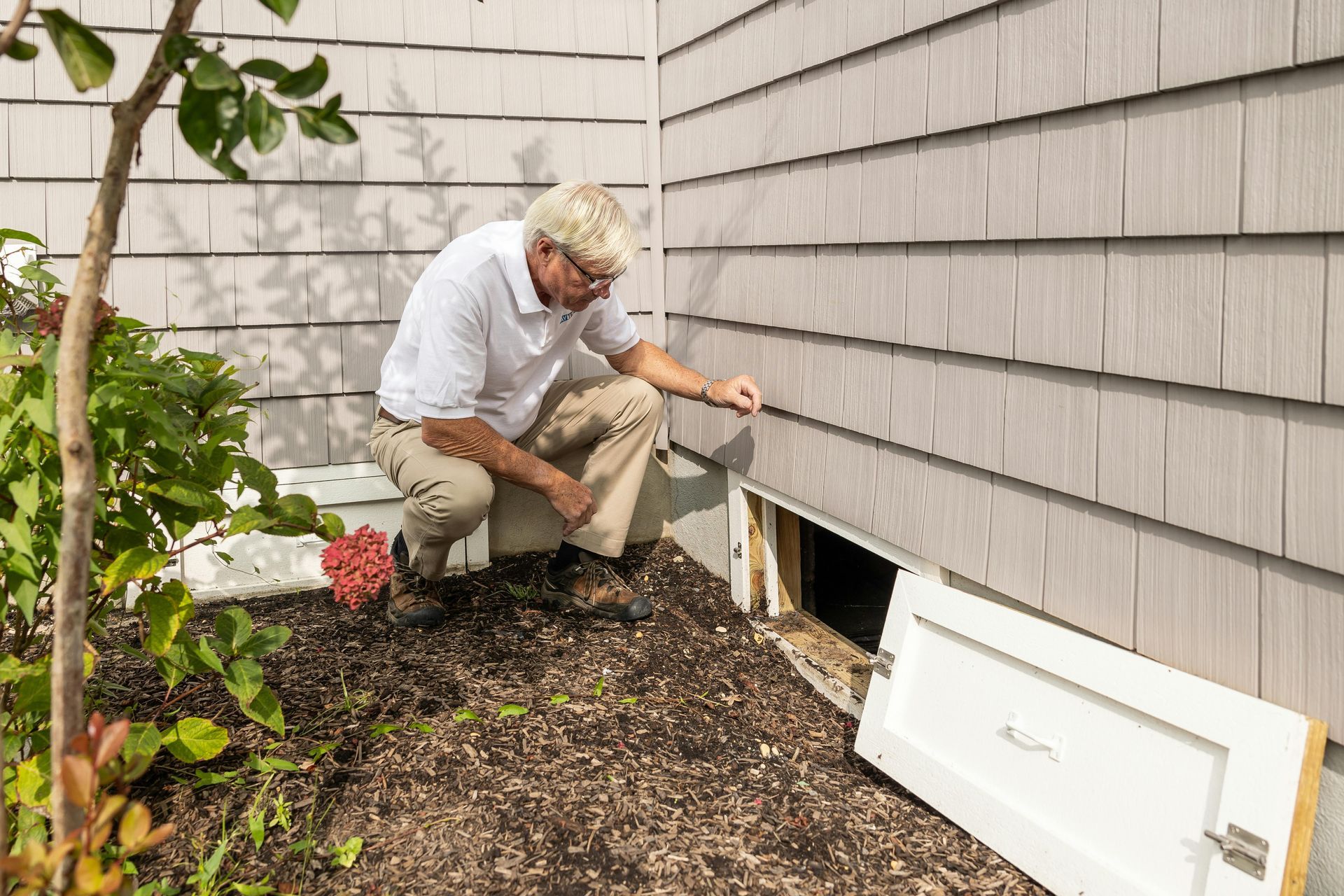 Person inspecting a crawlspace vent outside a house, beside white siding and mulch.