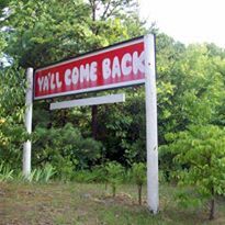A red and white sign that says `` ya 'll come back '' is surrounded by trees.