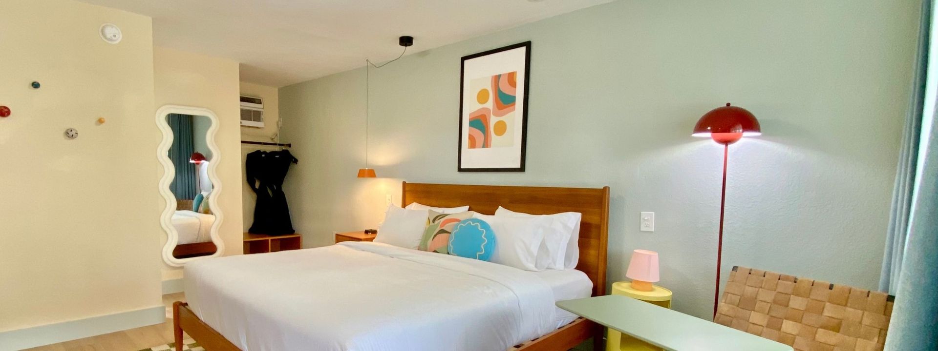 Bedroom with light wood furniture, patterned rug, and pastel colors; bed centered under yellow artwork.