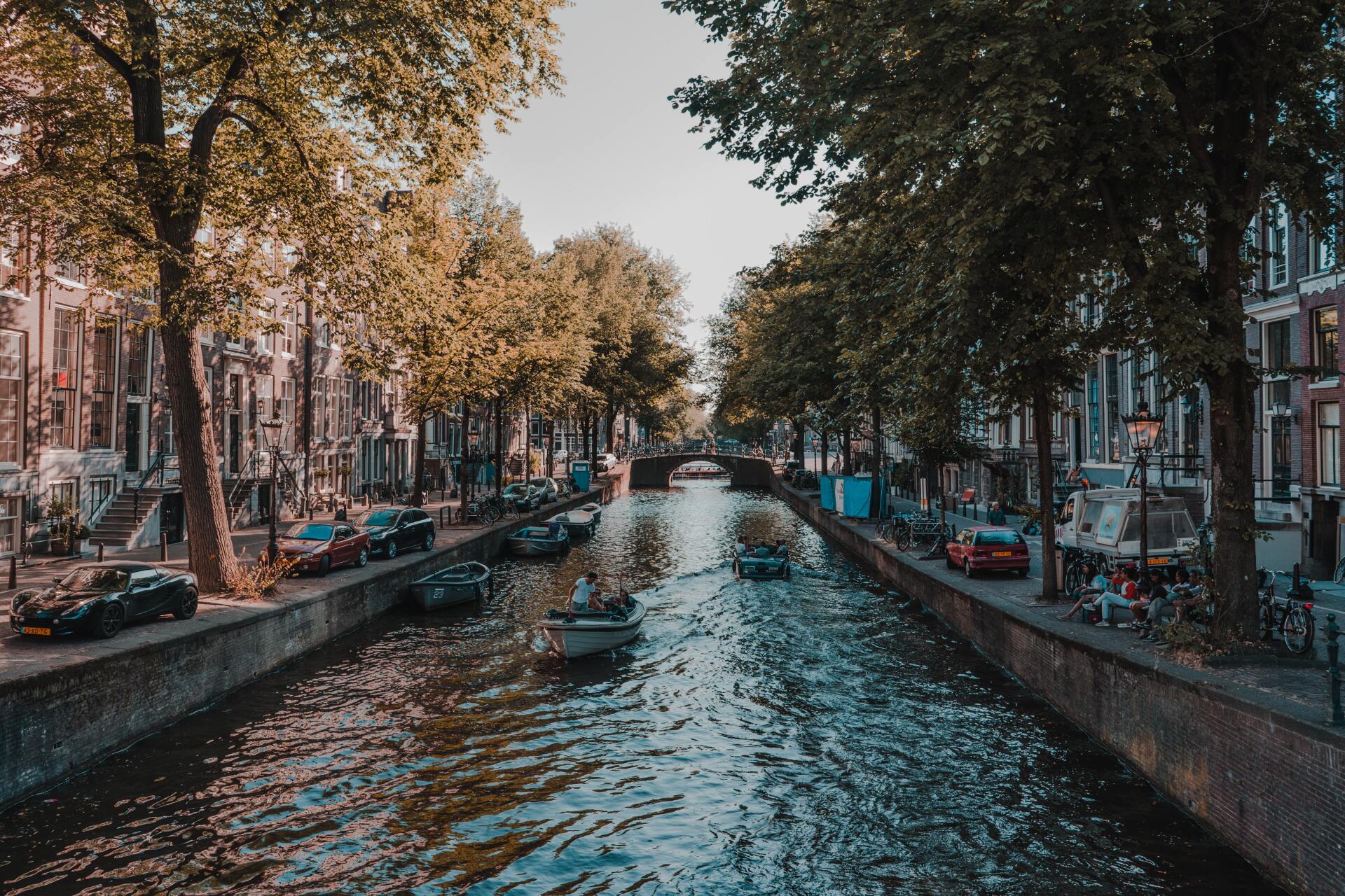 Canal in Amsterdam with boats, lined by buildings and trees; cars parked along the sides.