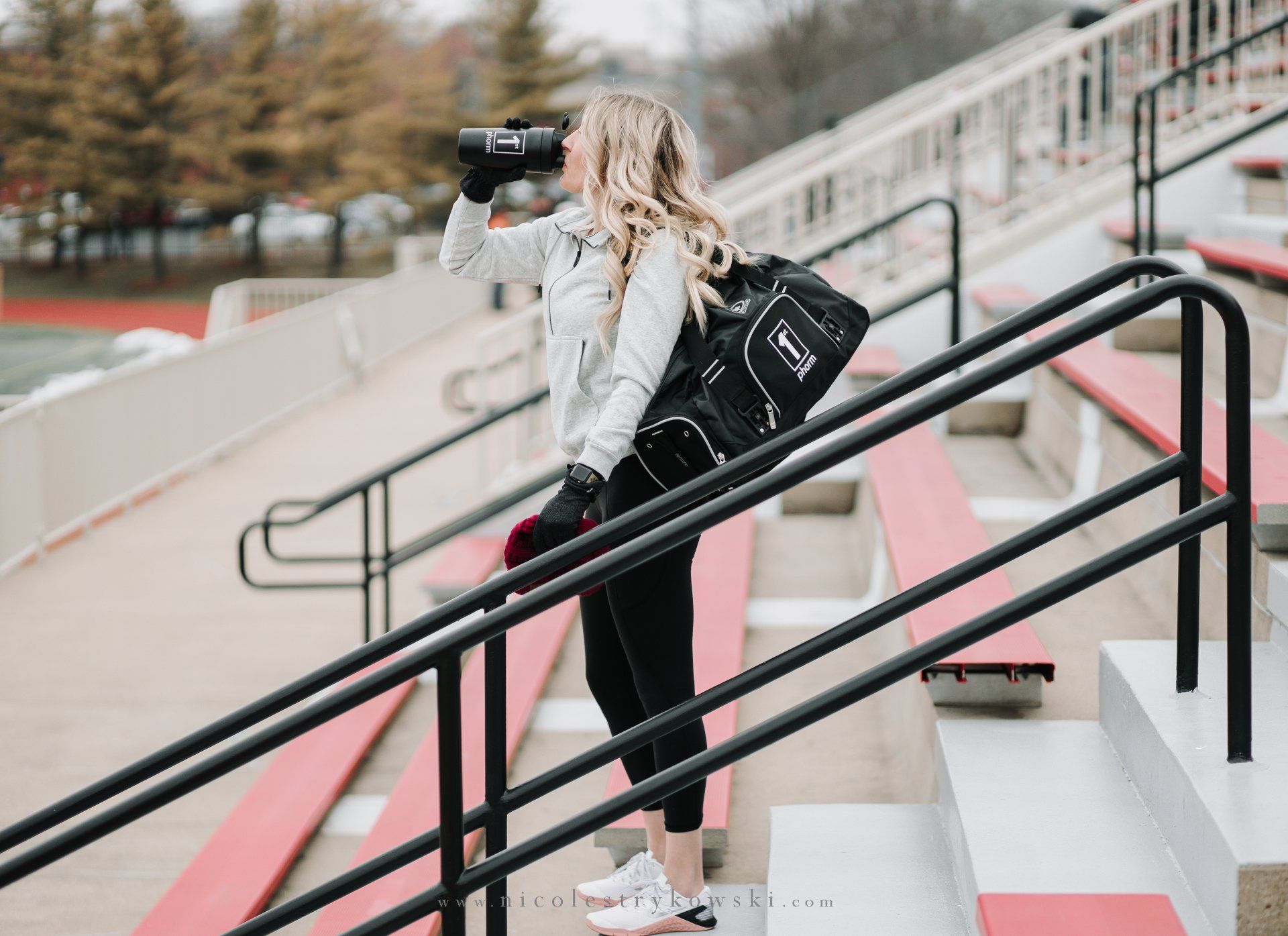 A woman is drinking water from a bottle while standing on stairs in a stadium.