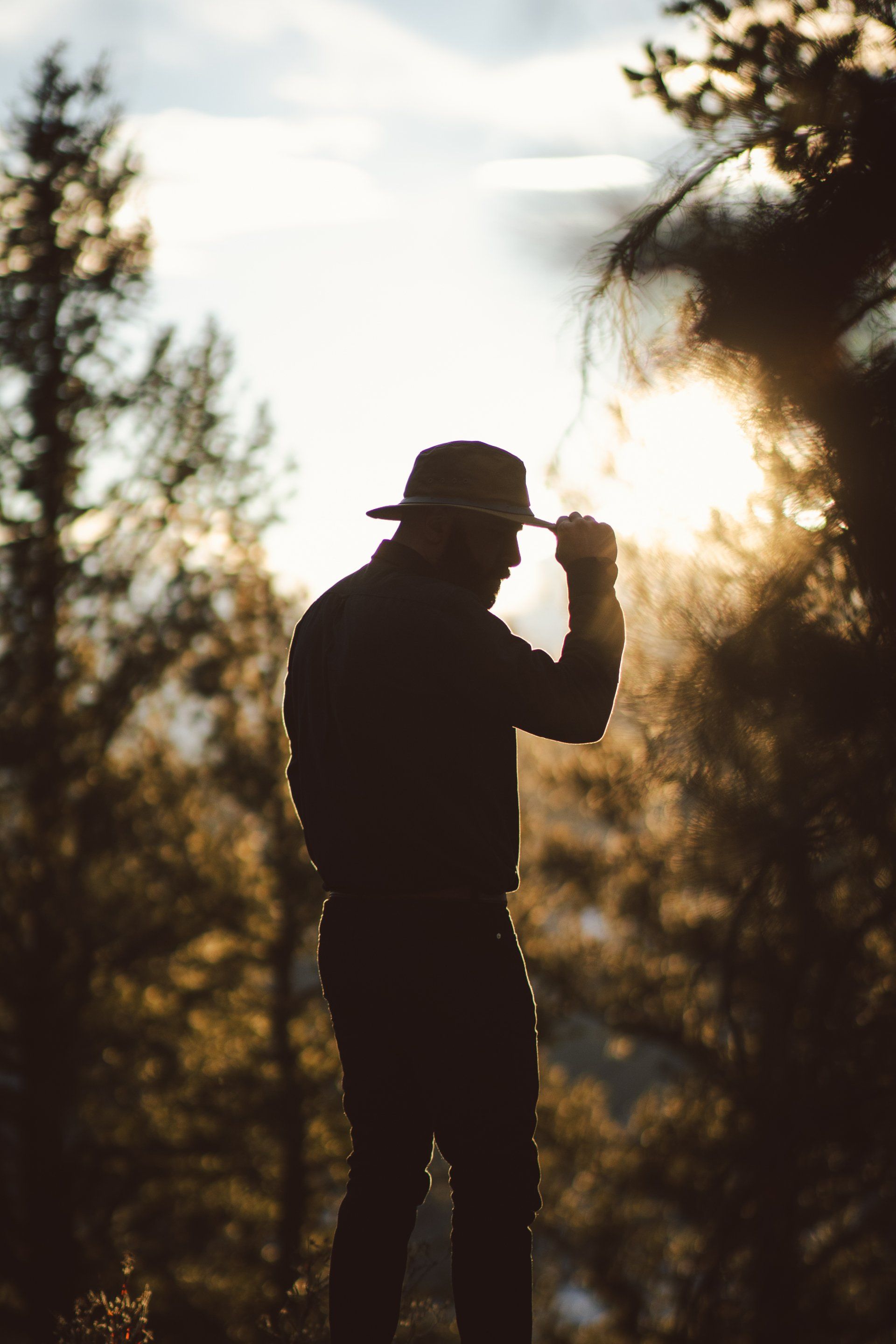 A man in a hat is standing in the woods at sunset.