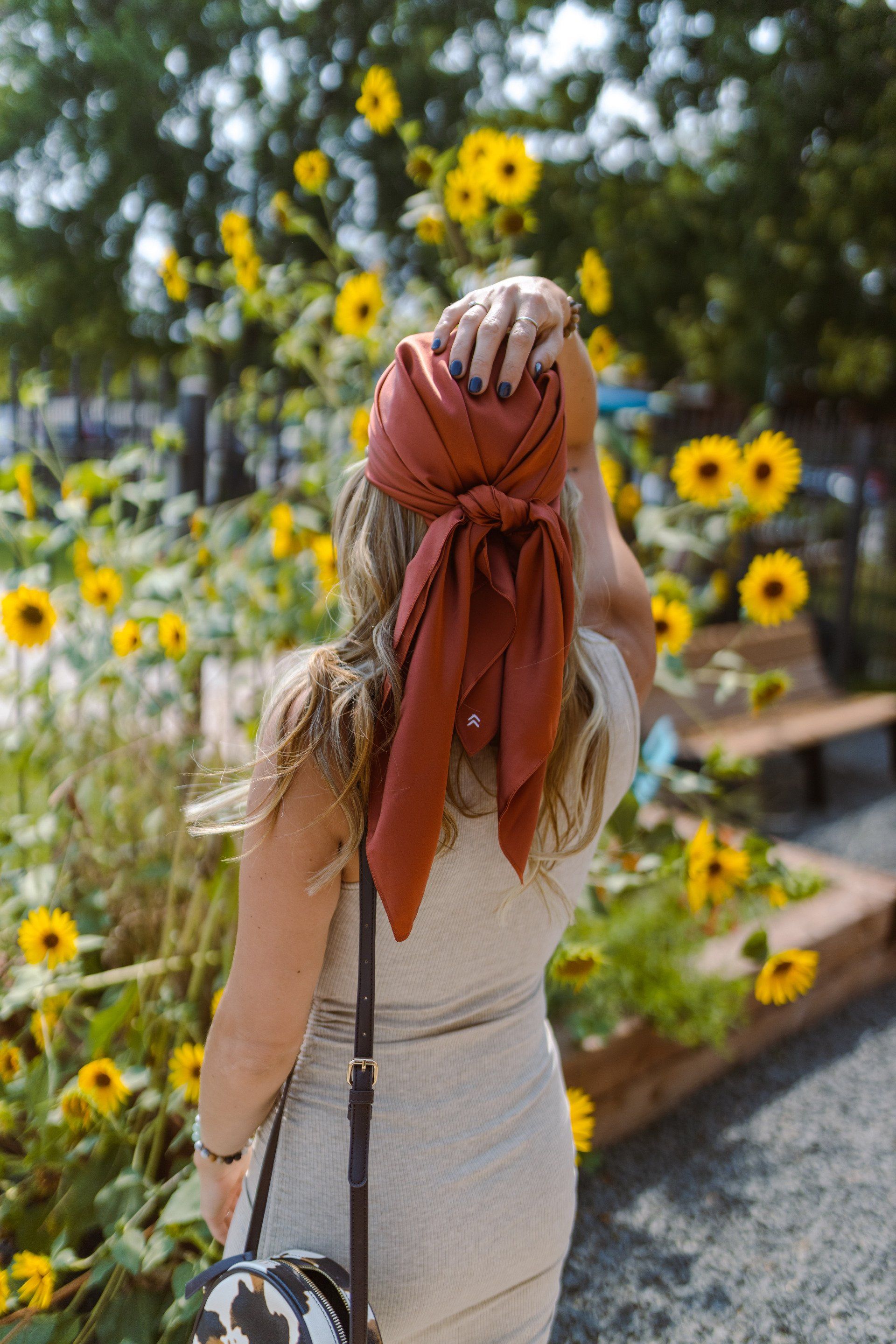 A woman wearing a scarf around her head is standing in front of a field of sunflowers.