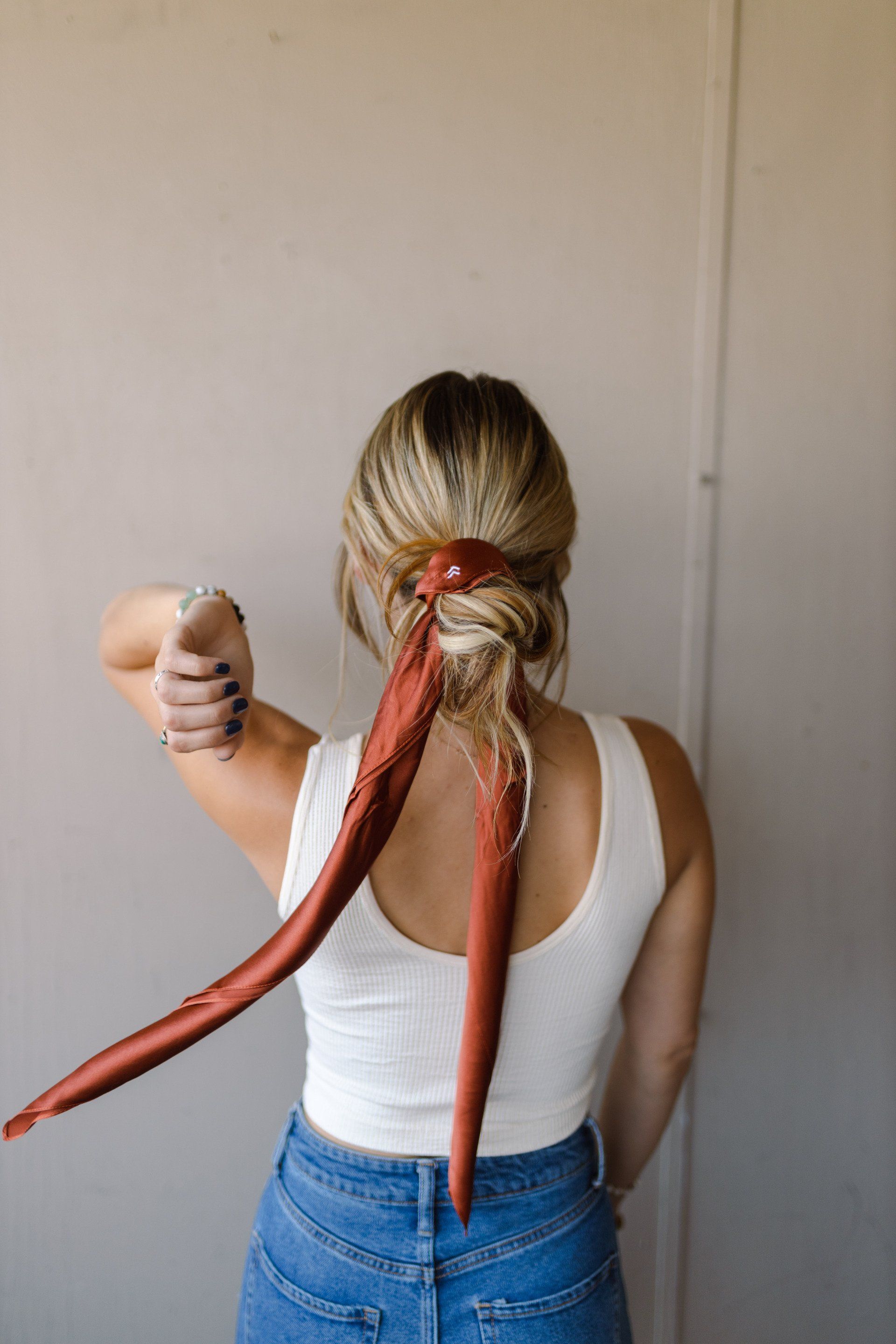 A woman in a white tank top and blue jeans is wearing a red scarf in her hair.