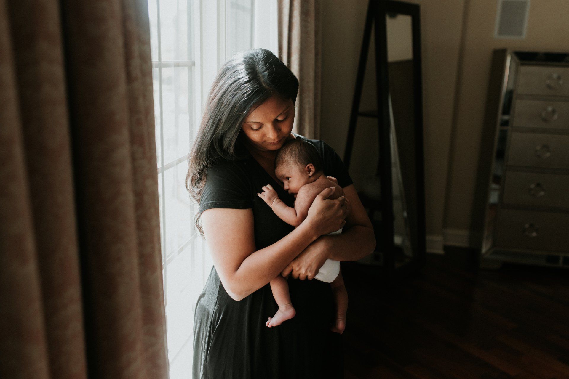 A woman is holding a baby in her arms in front of a window.