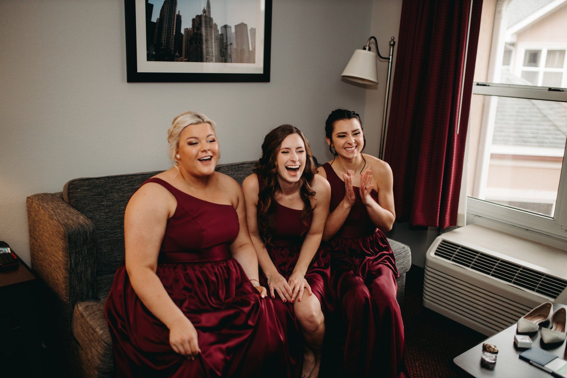 Three bridesmaids are sitting on a couch in a hotel room laughing.