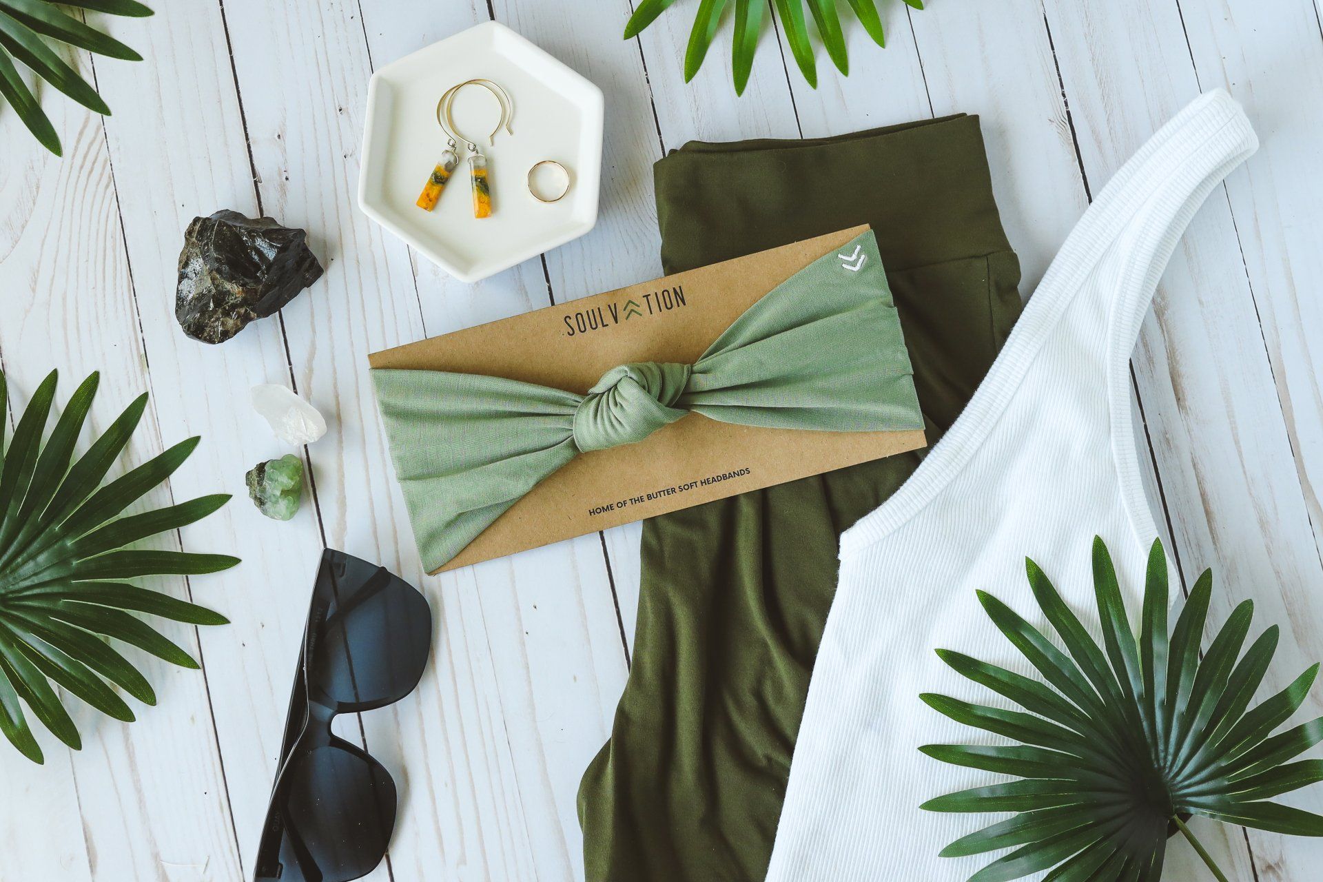A headband , sunglasses , earrings , and a white shirt are on a table surrounded by palm leaves.