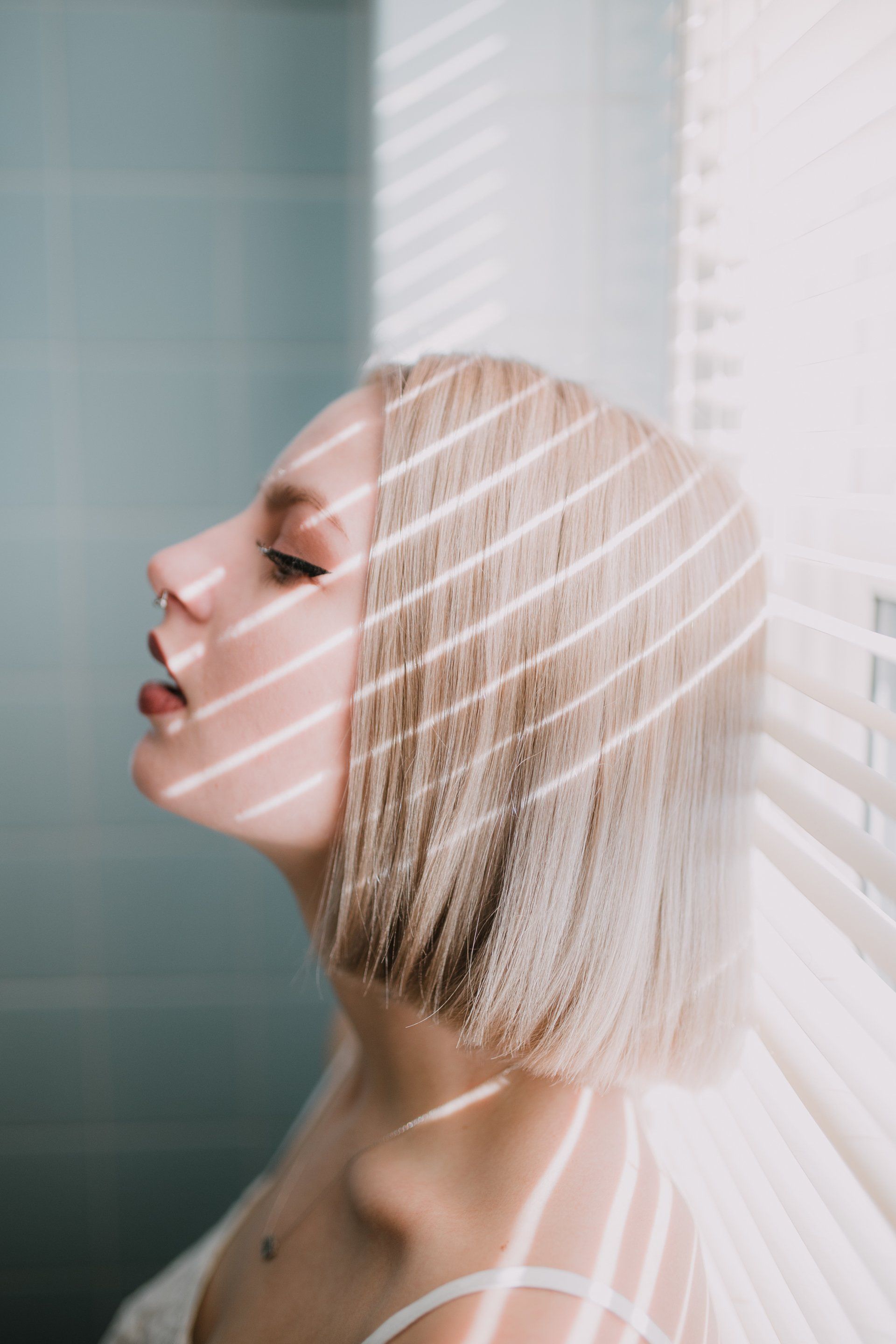 A woman with blonde hair is standing in front of a window with blinds.