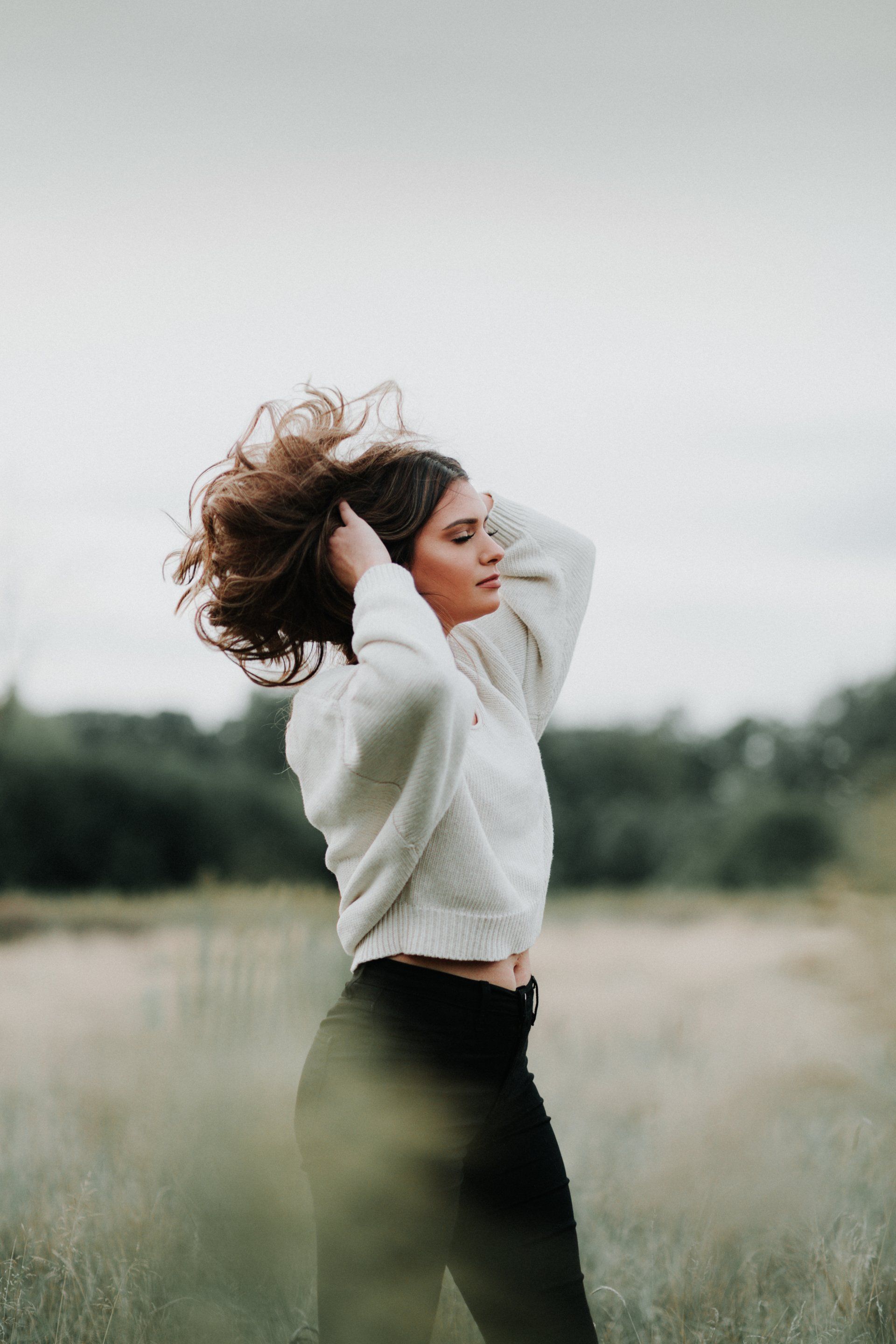 A woman in a white sweater is standing in a field with her hair blowing in the wind.
