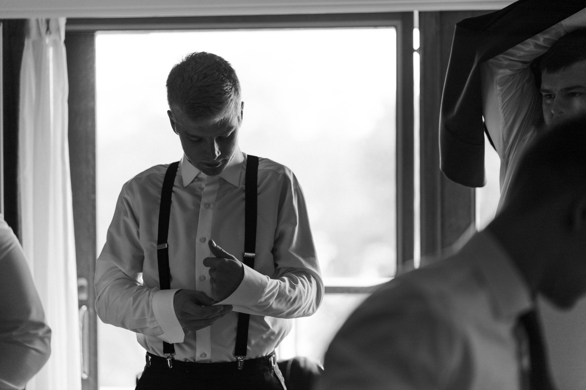 A man in a white shirt and suspenders is standing in front of a window.