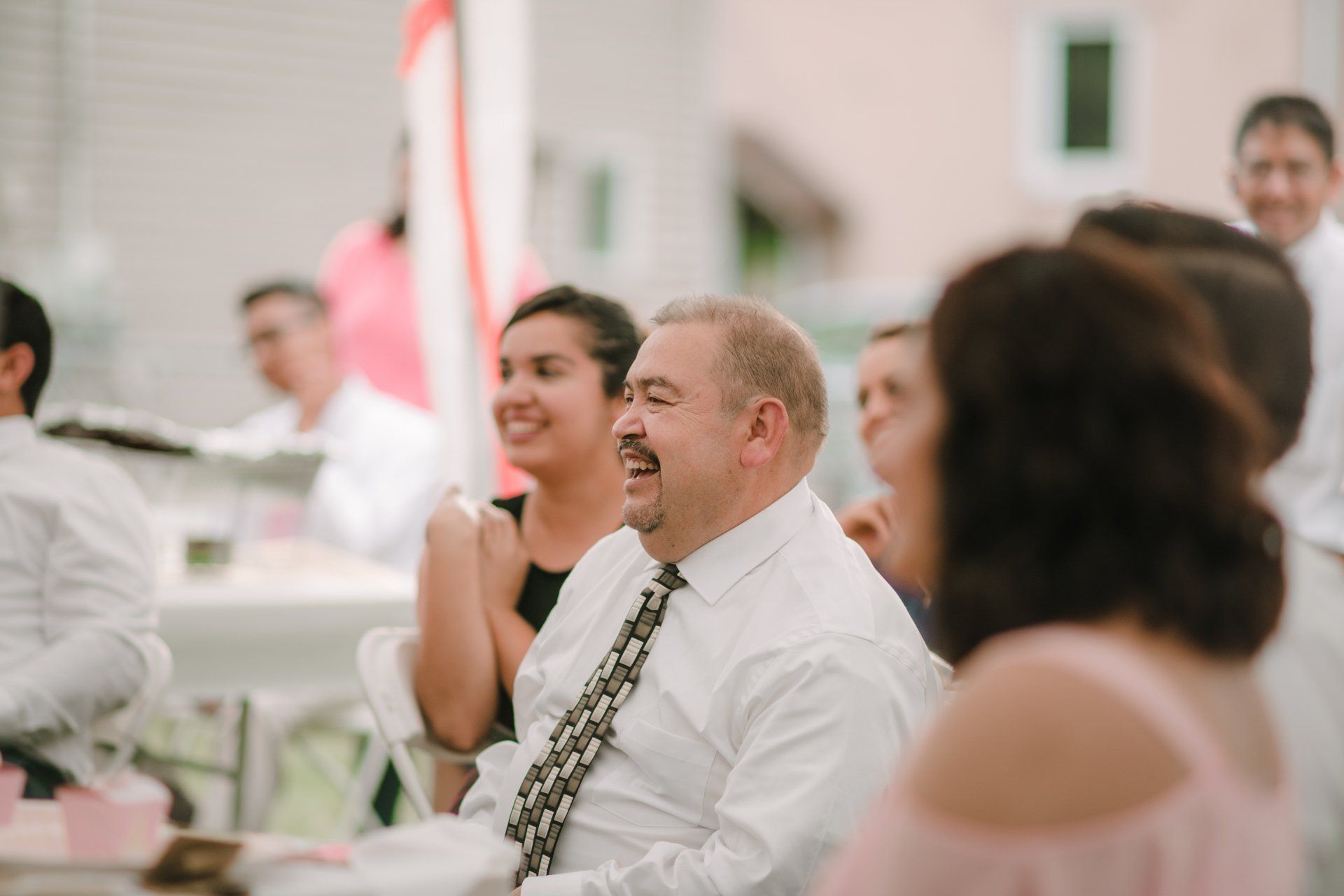 A man in a white shirt and tie is sitting at a table with other people at a wedding reception.