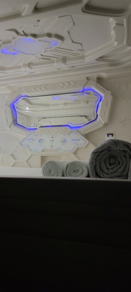 Interior of a futuristic capsule hotel room with geometric white panels, blue lighting, and rolled towels.