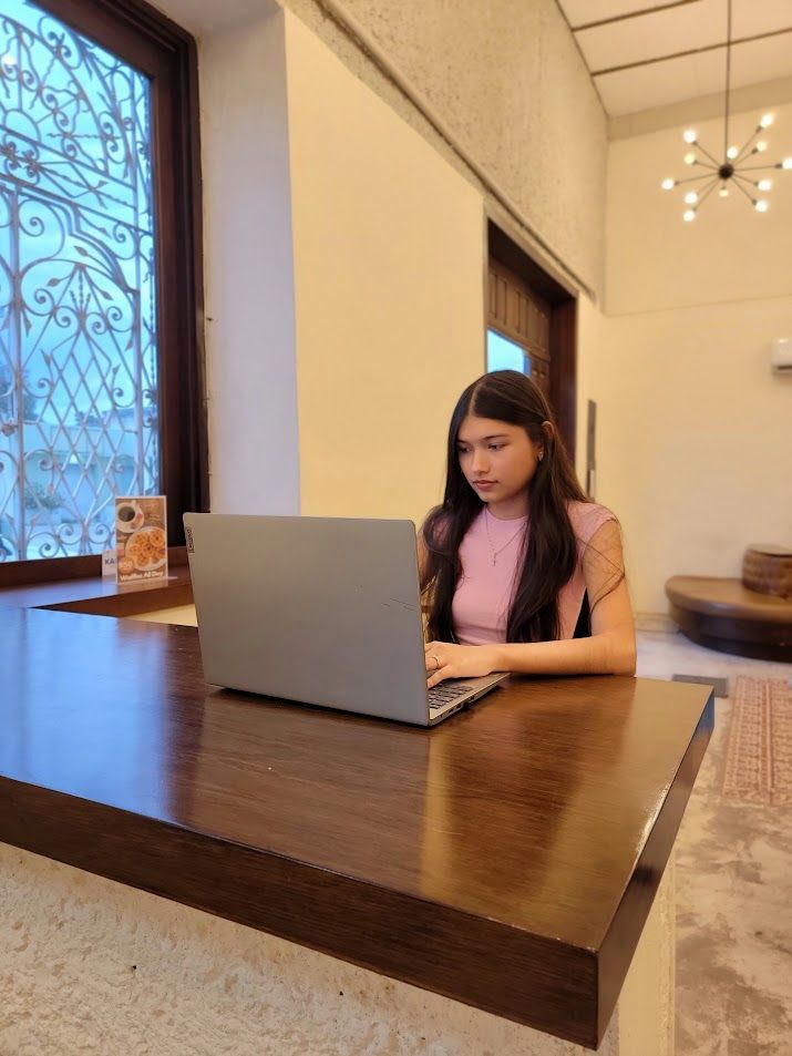 Woman using a laptop at a wooden counter near a window, focused expression. Cream walls and a light fixture in the background.