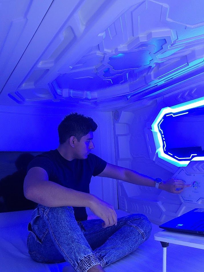 Man in futuristic pod, reaching toward a control panel. Blue lighting illuminates the interior.