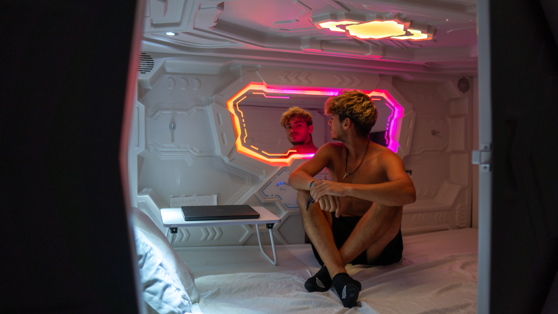 Man sitting in a futuristic pod-like room, looking at a mirror with neon lights.