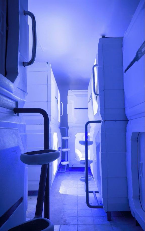 Capsule hotel room; stacked white sleeping pods in blue-lit hallway with handrails.