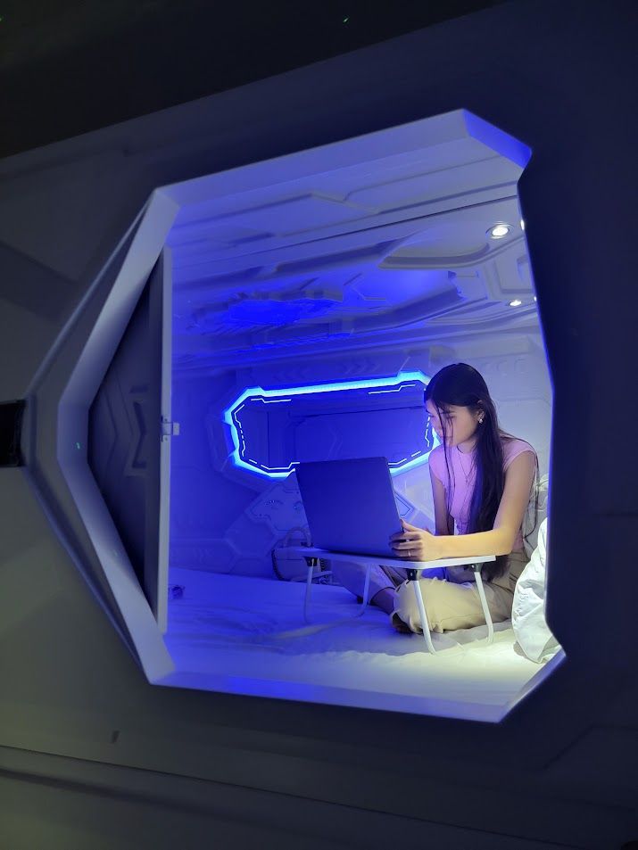 Woman working on a laptop inside a futuristic, lit-up sleep pod.